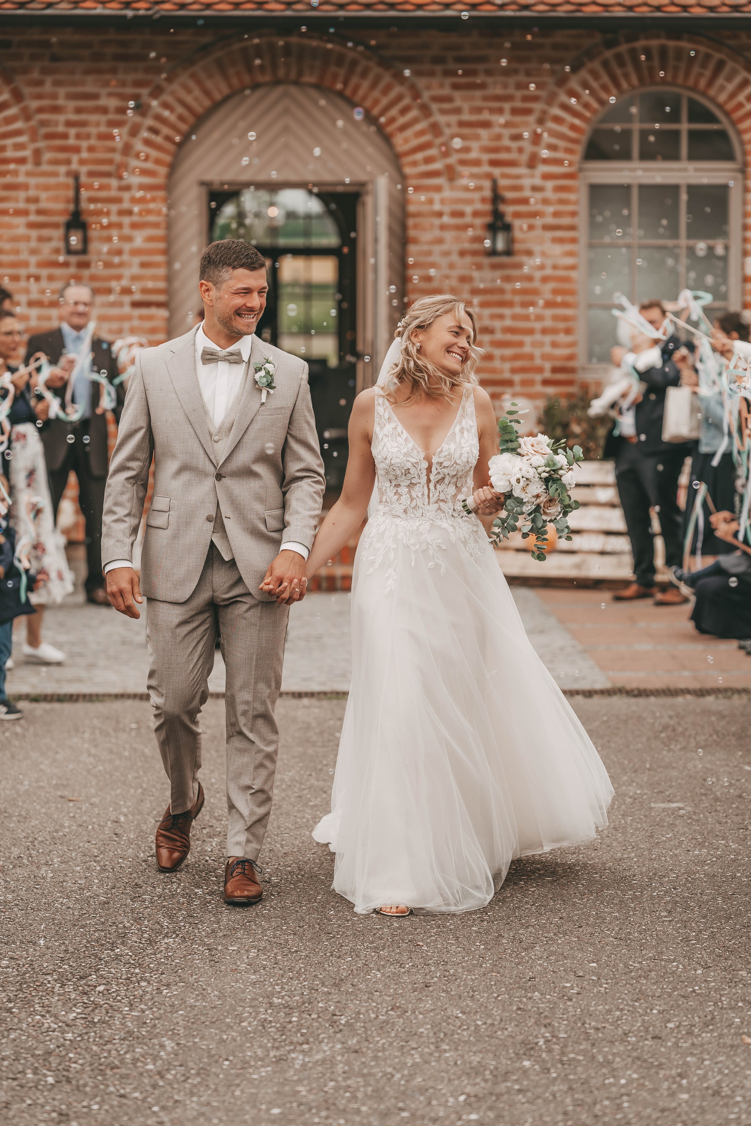 Ein Hochzeitspaar, ein Mann im grauen Anzug und eine Frau im weißen Brautkleid, halten Händchen und lächeln, während sie aus einem Backsteingebäude gehen. Im Hintergrund stehen Gäste, die Jubeln und Seifenblasen pusten.