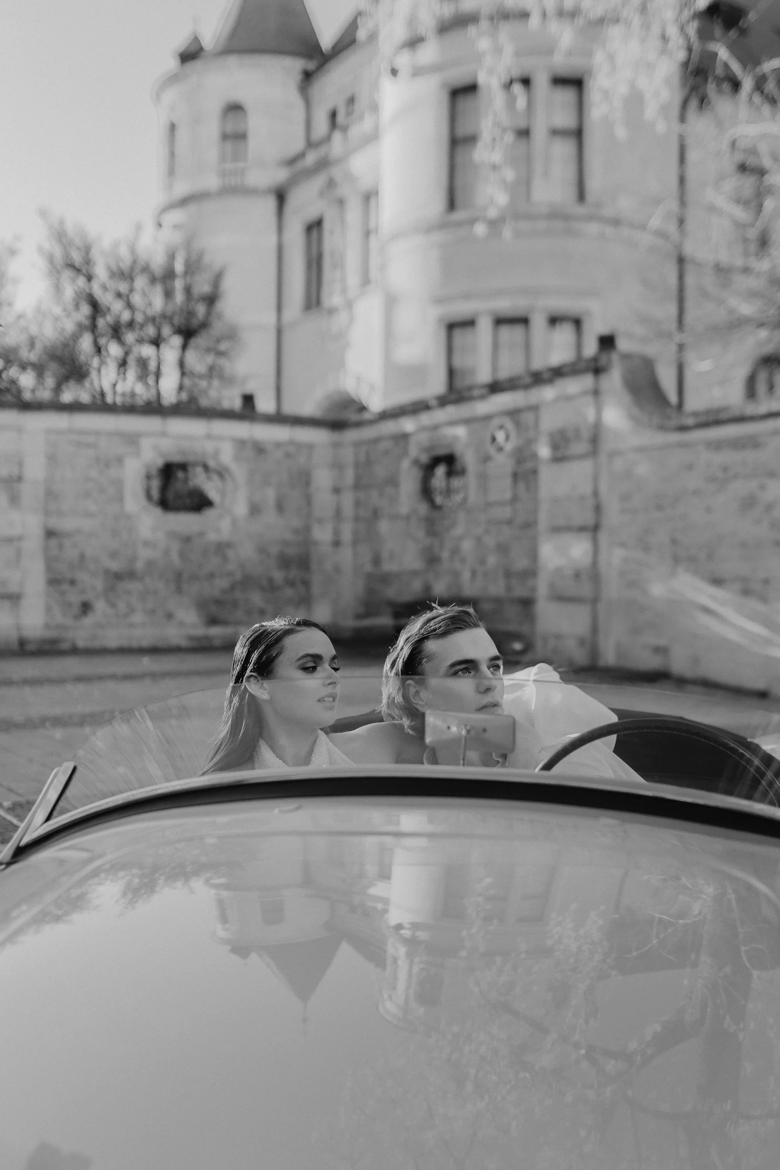 Zwei junge Menschen sitzen in einem Cabrio, im Hintergrund ein historisches Gebäude. Schwarz-Weiß-Fotografie.