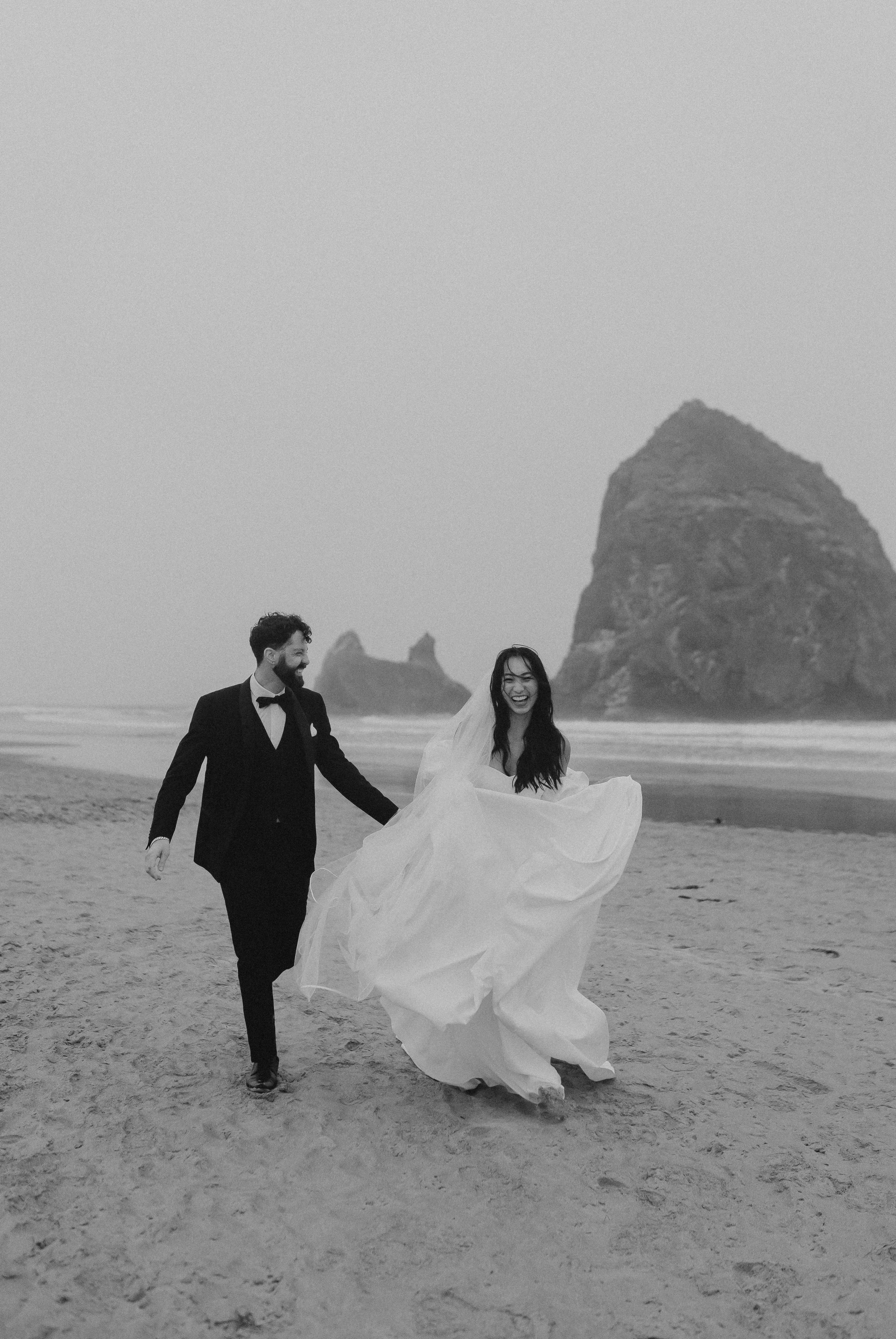 Ein lachendes Brautpaar in Hochzeitskleidung, das an einem Strand spaziert. Im Hintergrund sind Felsen und das Meer sichtbar, in schwarz-weiß.