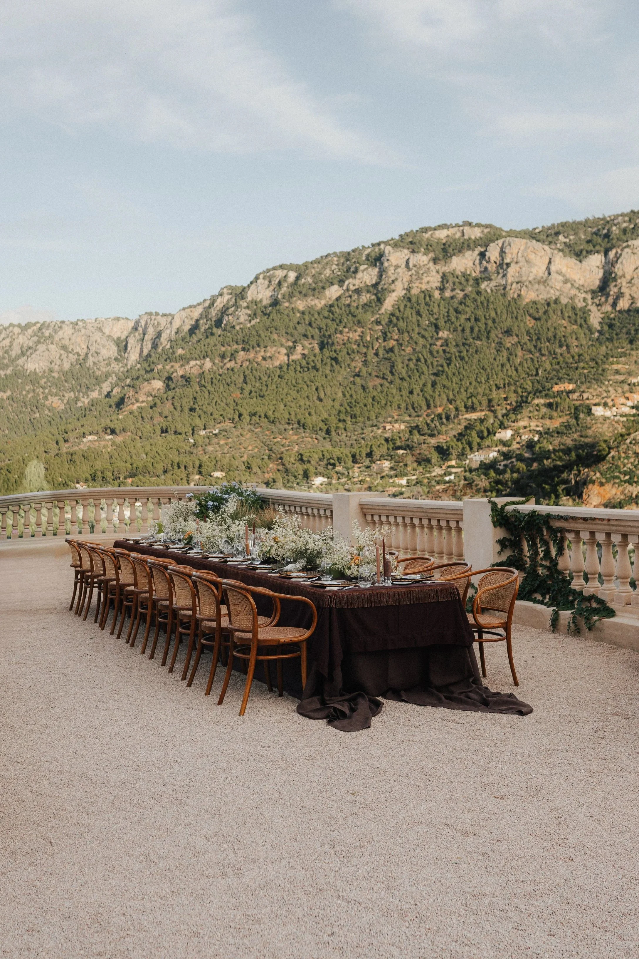 Ein langer festlicher Esstisch mit dunkelbraunem Tuch, gedeckt für ein Abendessen, auf einer Terrasse mit Blick auf Berge.