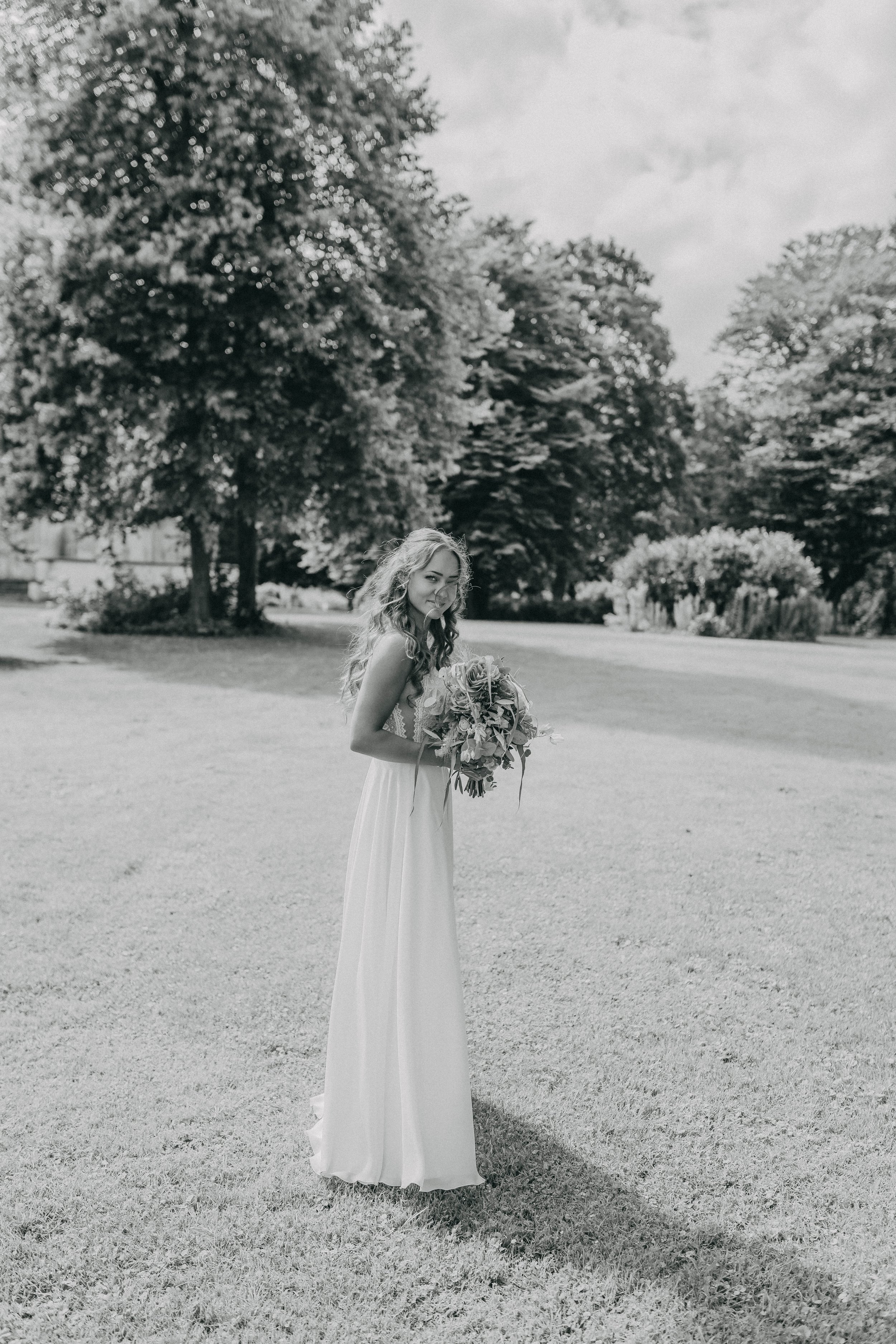 Frau im Hochzeitskleid mit Blumenstrauß auf einer Wiese, umgeben von Bäumen.