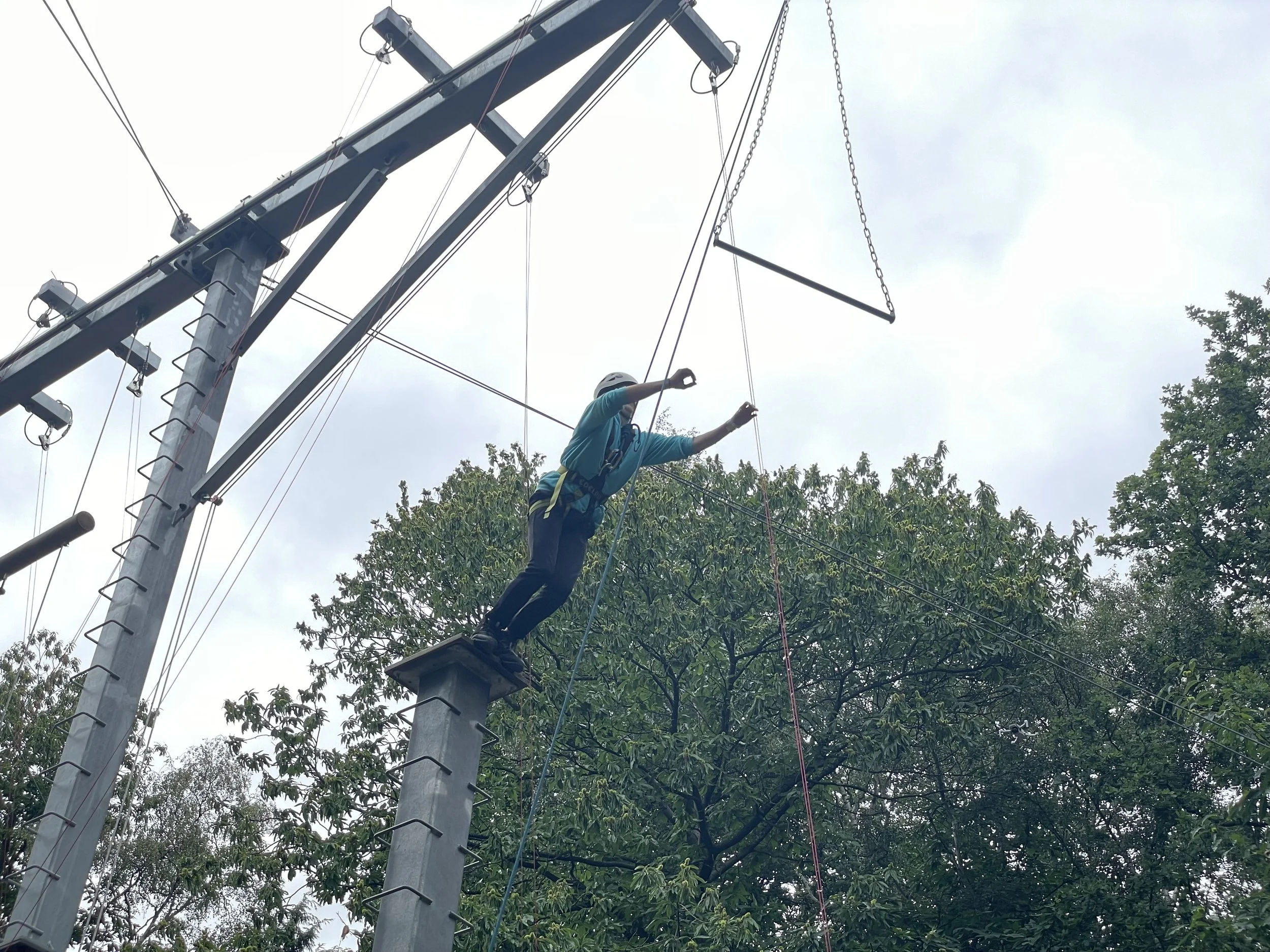 A person wearing a helmet and harness jumping on a high ropes course on an elevated wooden platform among tall trees.