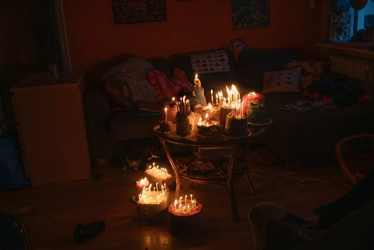 Living room with birthday cakes and lit candles on tables and in front of the couches, with cushions and a cat on the floor.