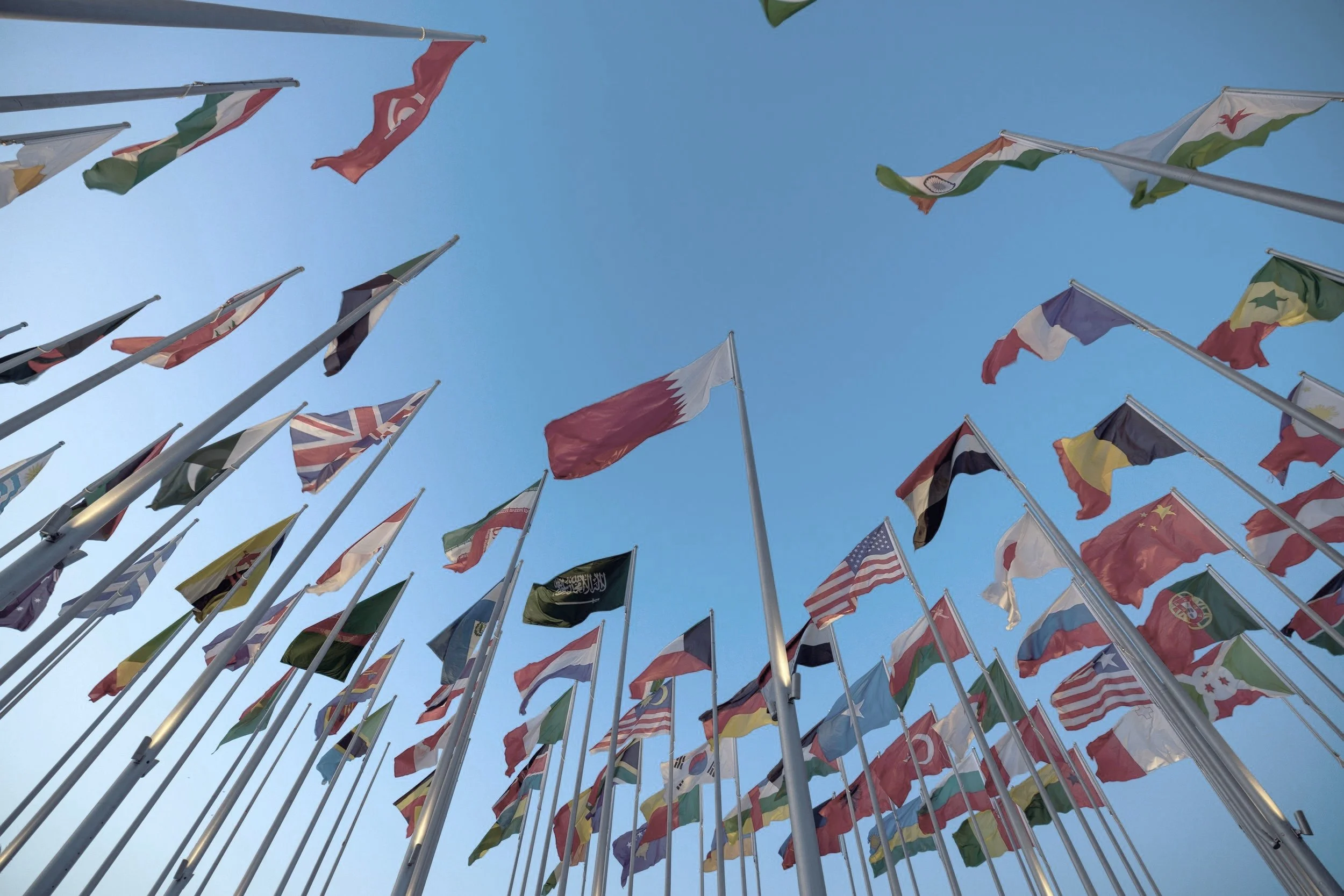 A low-angle view of multiple flags from various countries flying on flagpoles against a clear blue sky.