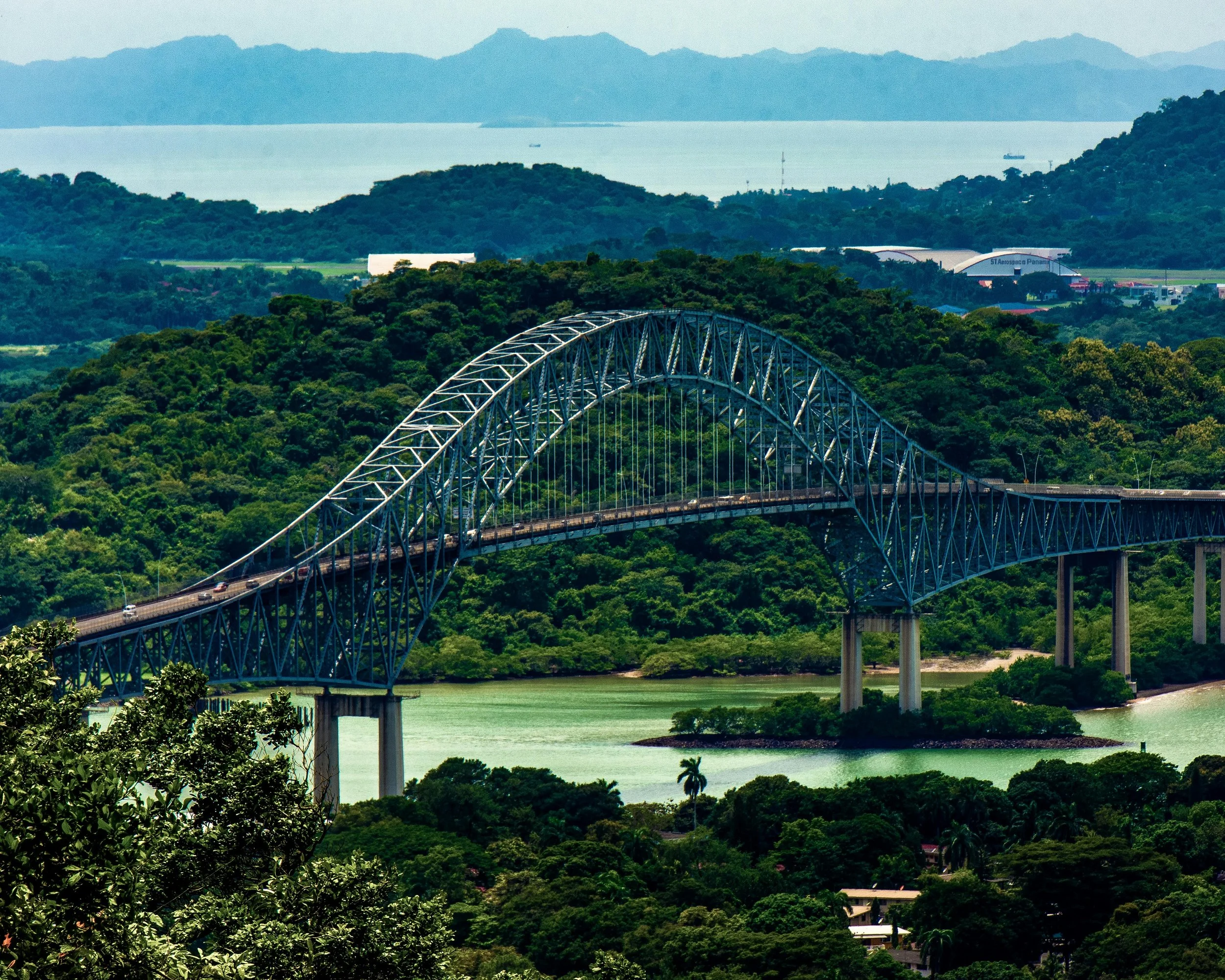 A scenic view of a bridge crossing over a river surrounded by lush green trees, with a mountain range in the background and a bay with boats.