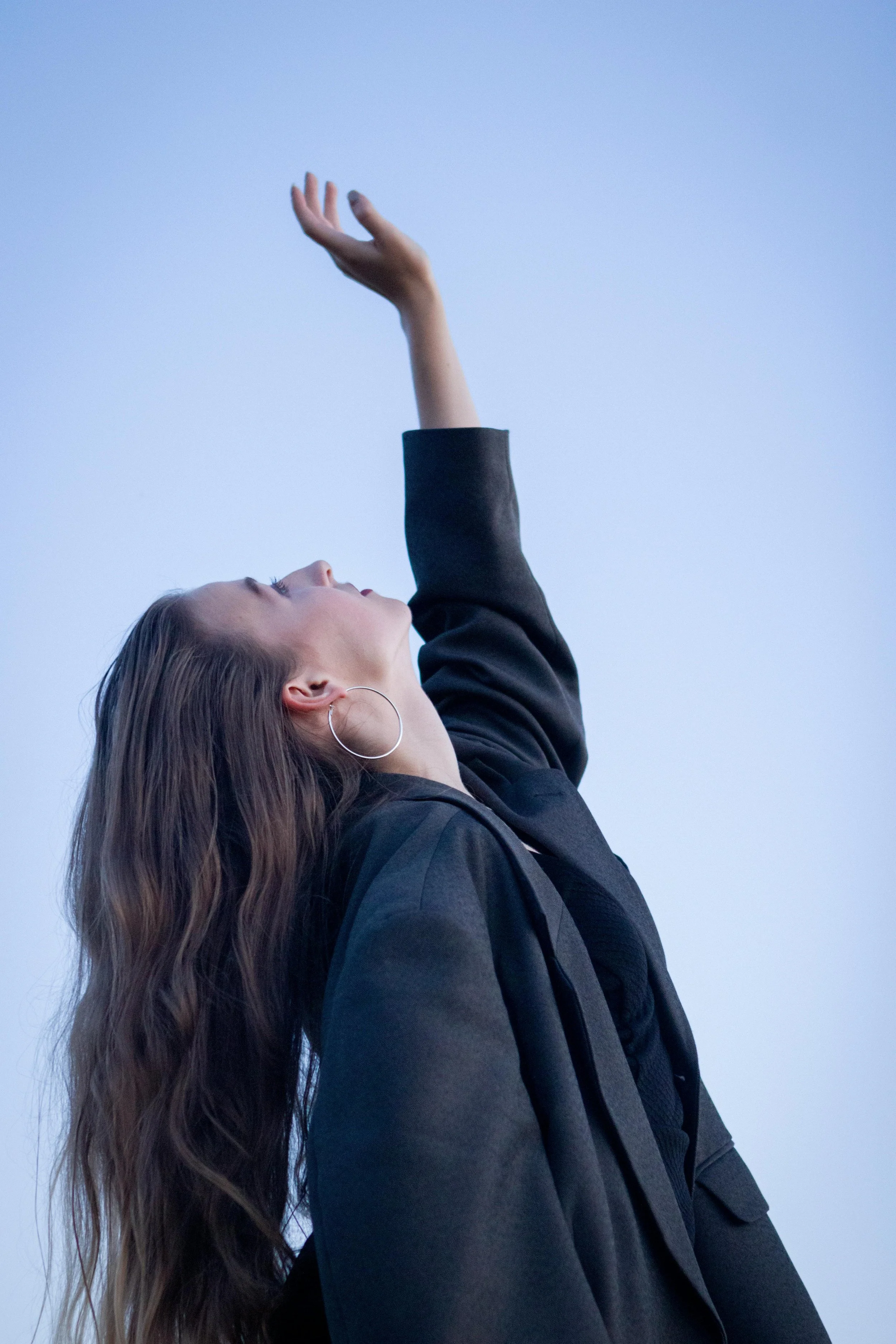 A woman with long wavy brown hair, wearing a dark blazer, is looking up with her eyes closed and one arm raised, set against a clear blue sky.