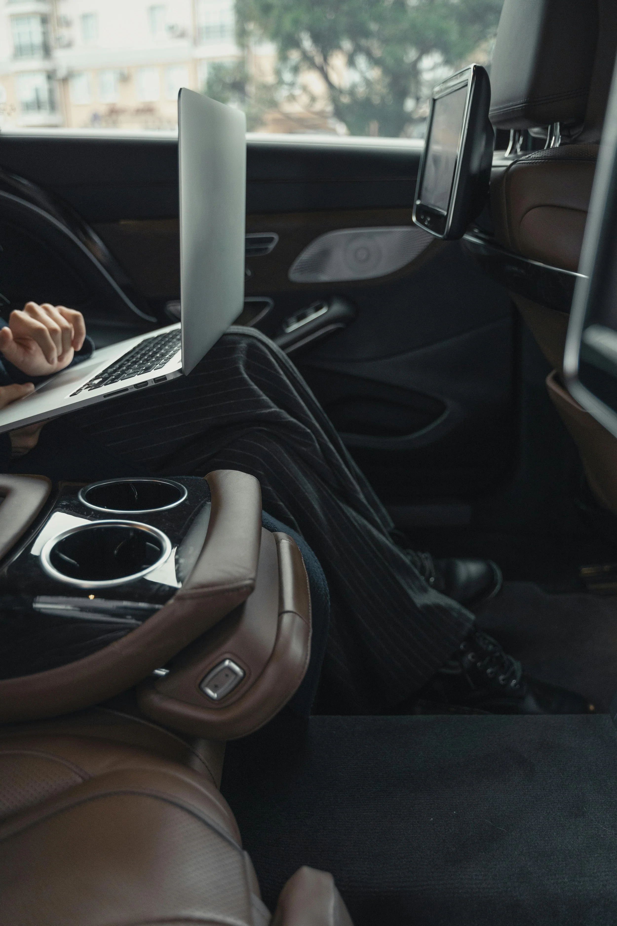 Inside a vehicle showing a person's lap, a laptop on their legs, a brown leather seat with built-in cup holders, and multiple screens attached to the headrests of the front seats