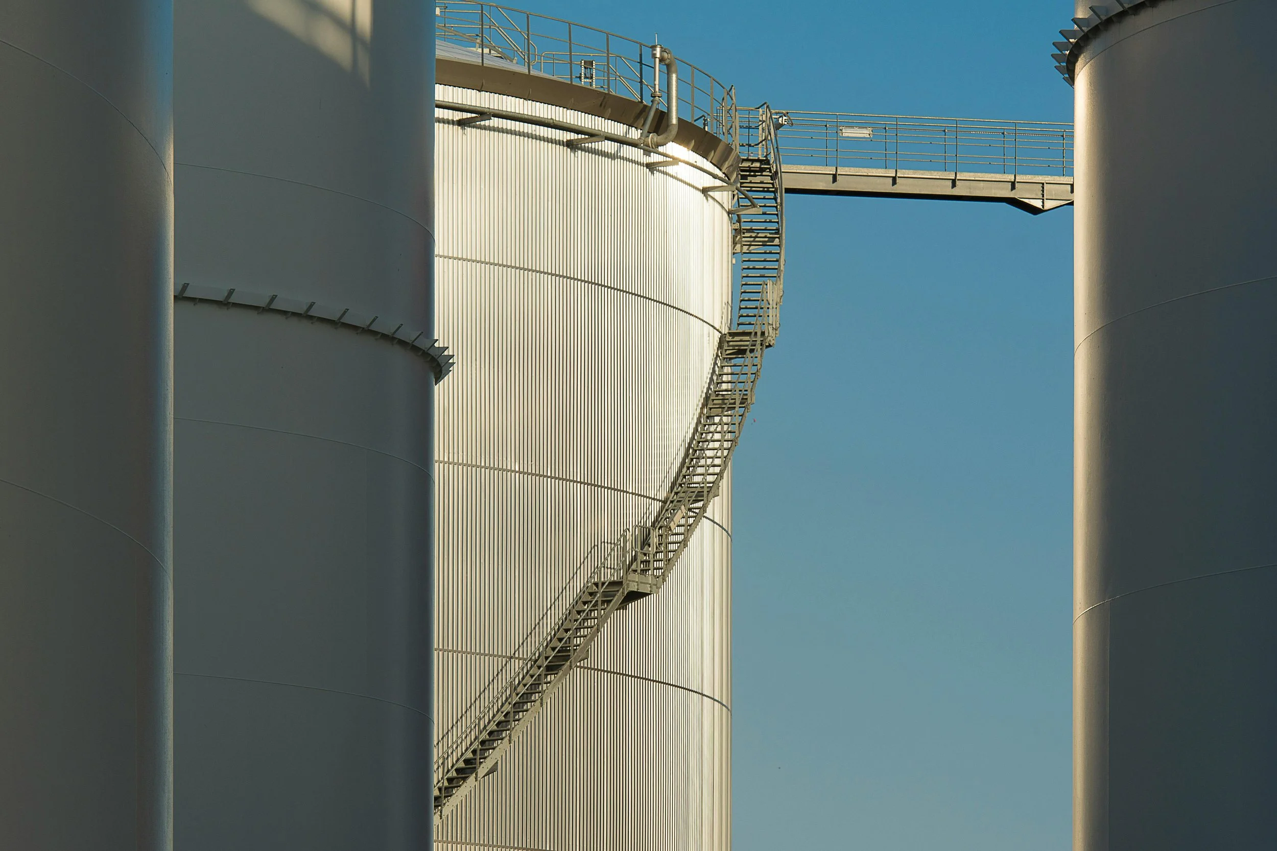 Large industrial storage tanks with metal staircase and railing, under a clear blue sky.