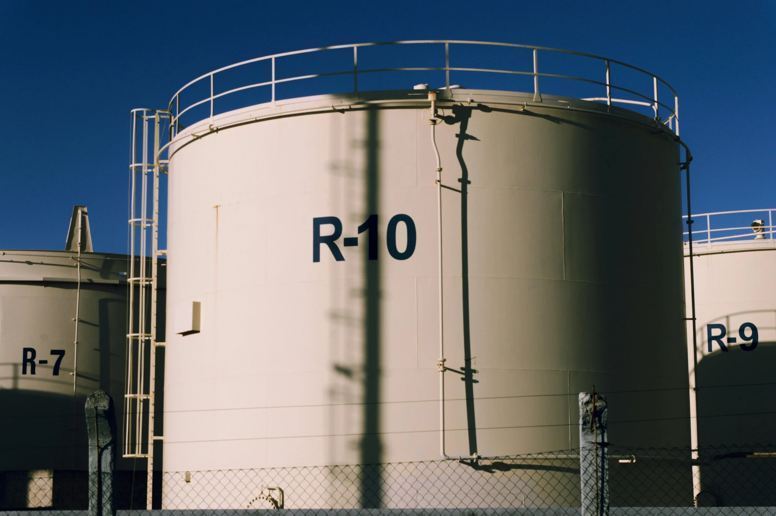 Large white industrial storage tanks with black markings R-10 and R-9, surrounded by chain-link fence, under a clear blue sky.