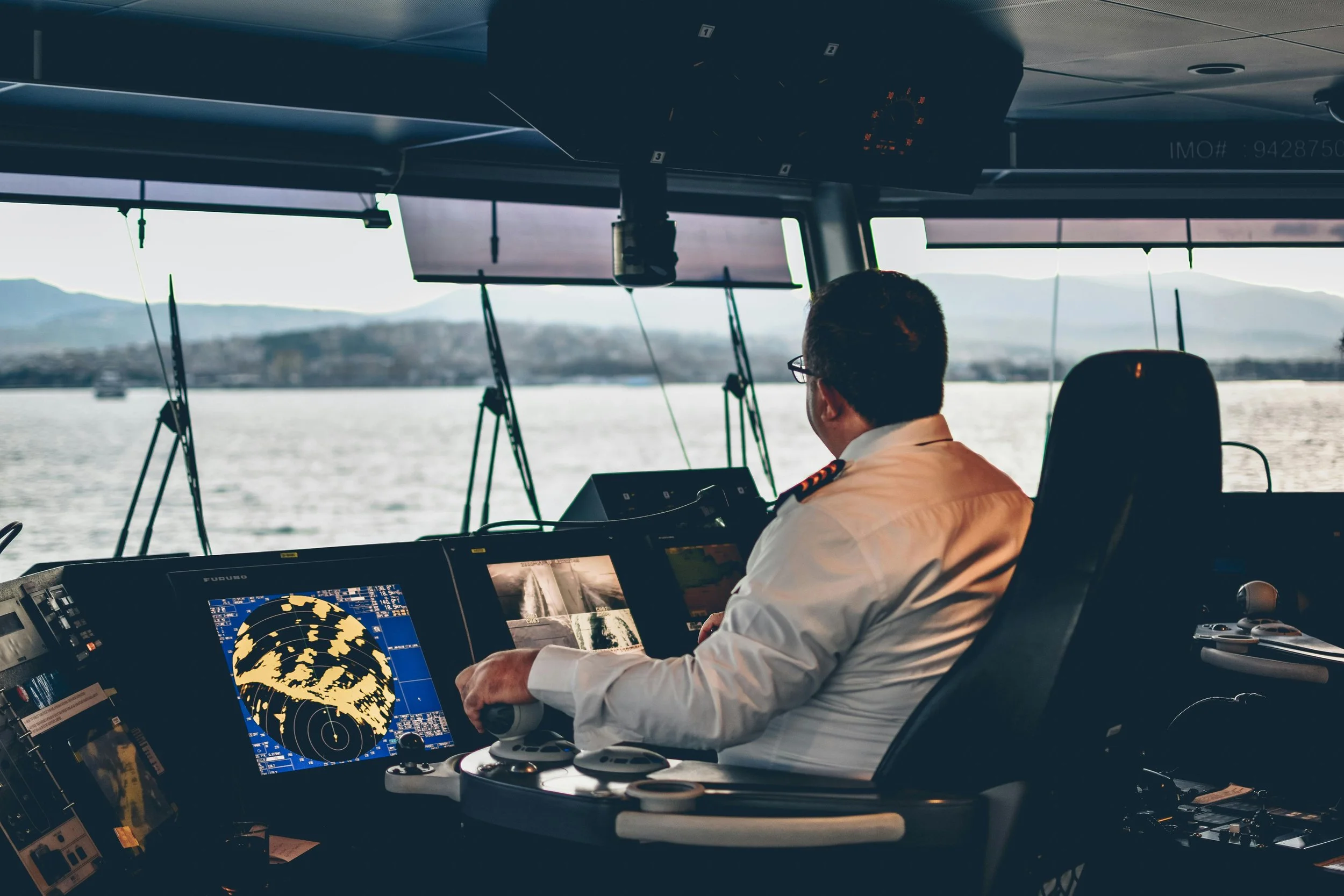 A maritime vessel captain navigating through waters with electronic navigation controls and screens, with a scenic view of water and distant land outside the window.
