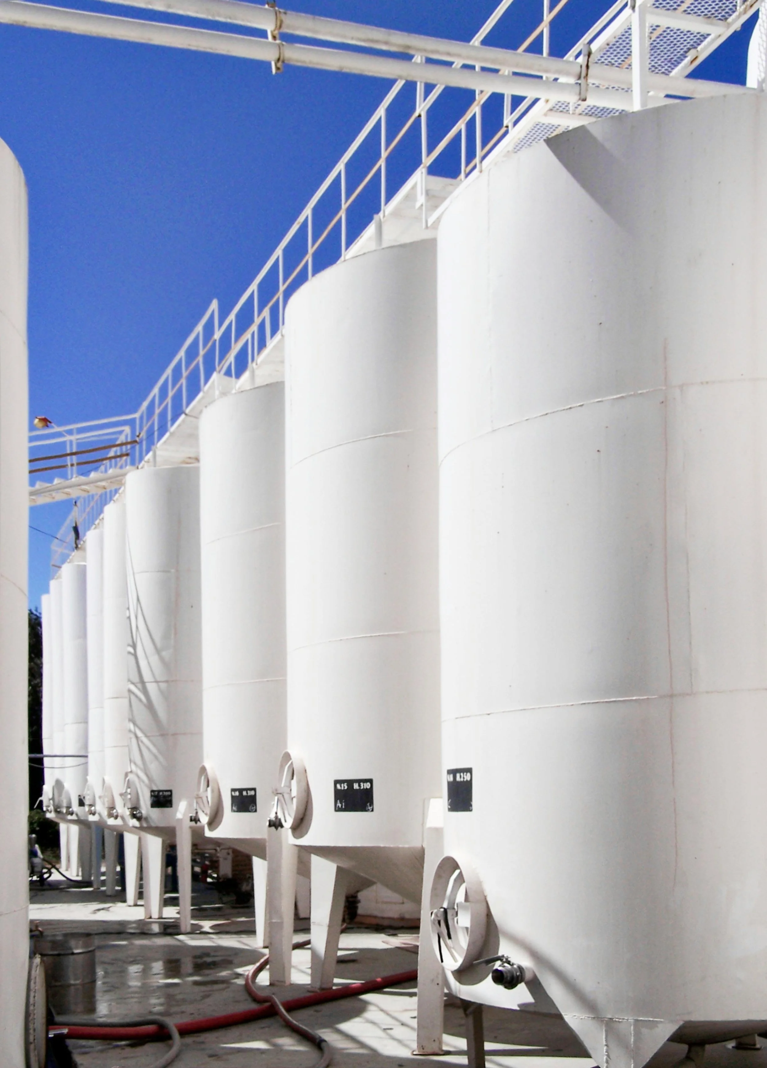A row of large white tanks or silos with metal railings and small labels at the bottom, under a bright blue sky.