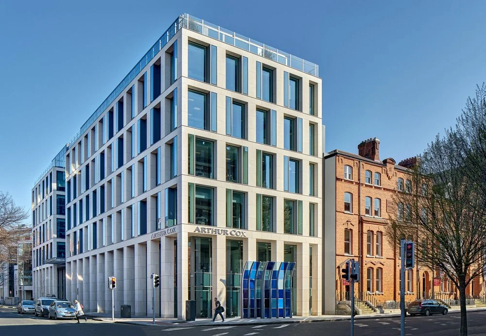 Modern multi-story office building with glass windows and white framing, located on a city street corner with cars and pedestrians, compared to older brick buildings nearby.