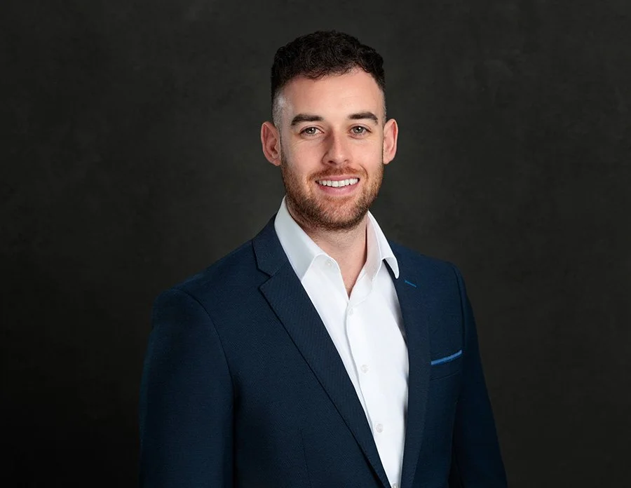 A young man with dark curly hair, beard, and blue eyes, wearing a navy blue suit and white dress shirt, smiling against a dark gray background.