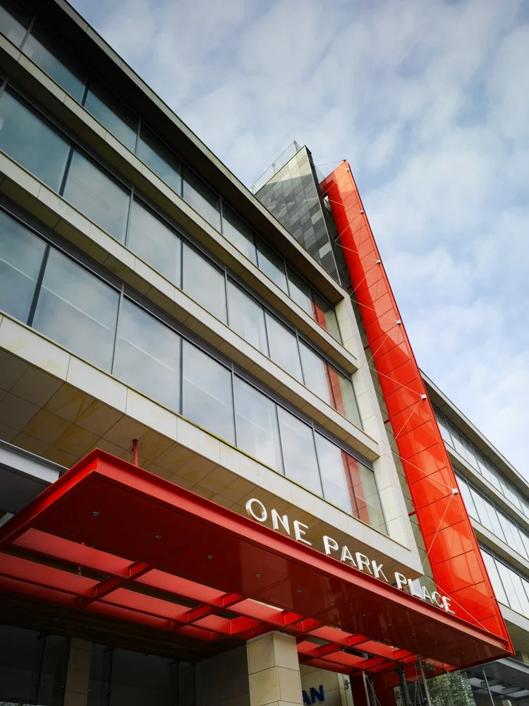 Modern multi-story building with large glass windows and red architectural accents, including a large red vertical element and a red canopy at the entrance, with the sign 'ONE PARK PLACE' on the building.