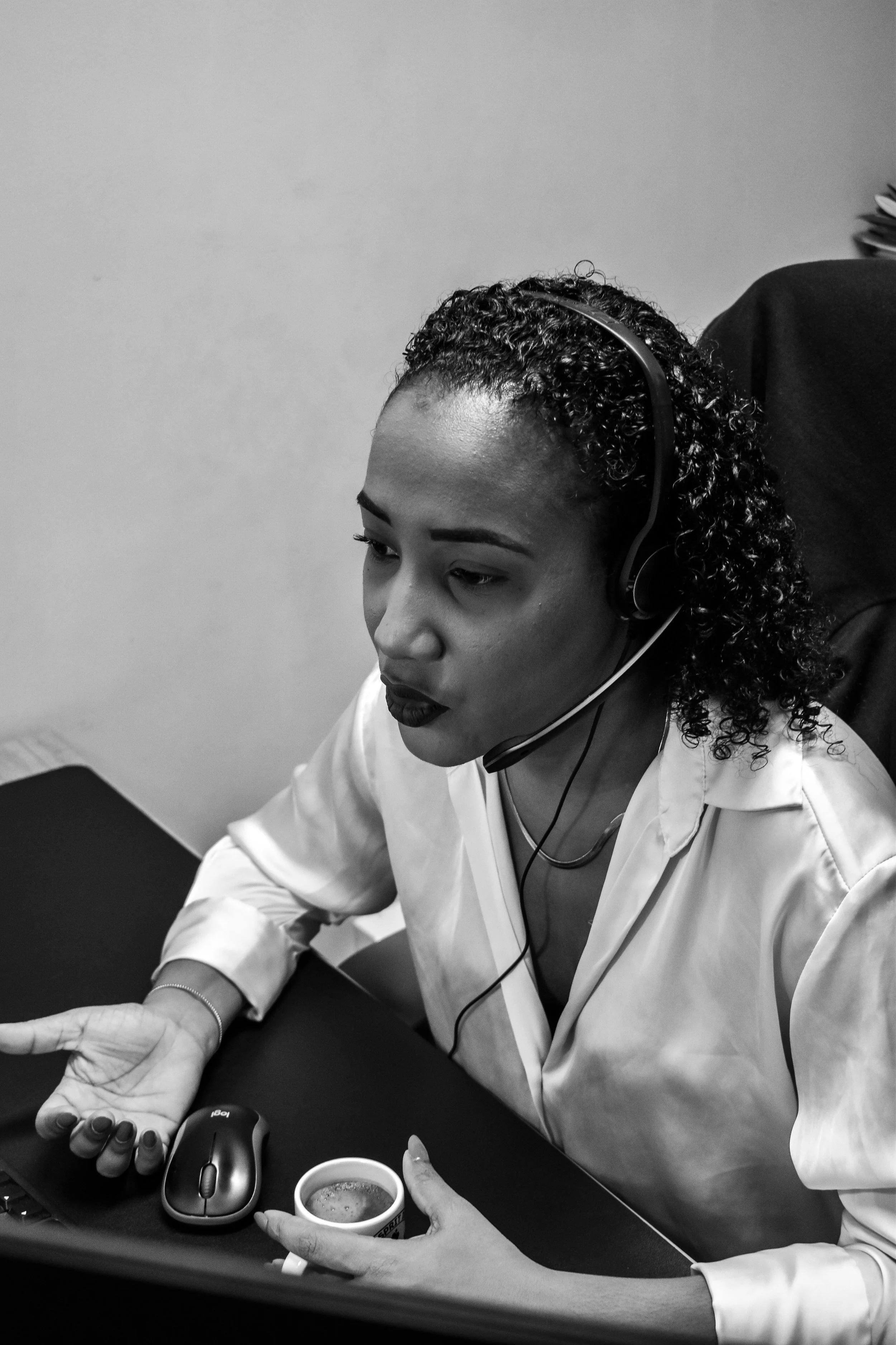 Femme portant un casque avec microphone, assise devant un ordinateur, tenant une tasse, en conversation ou en travail.