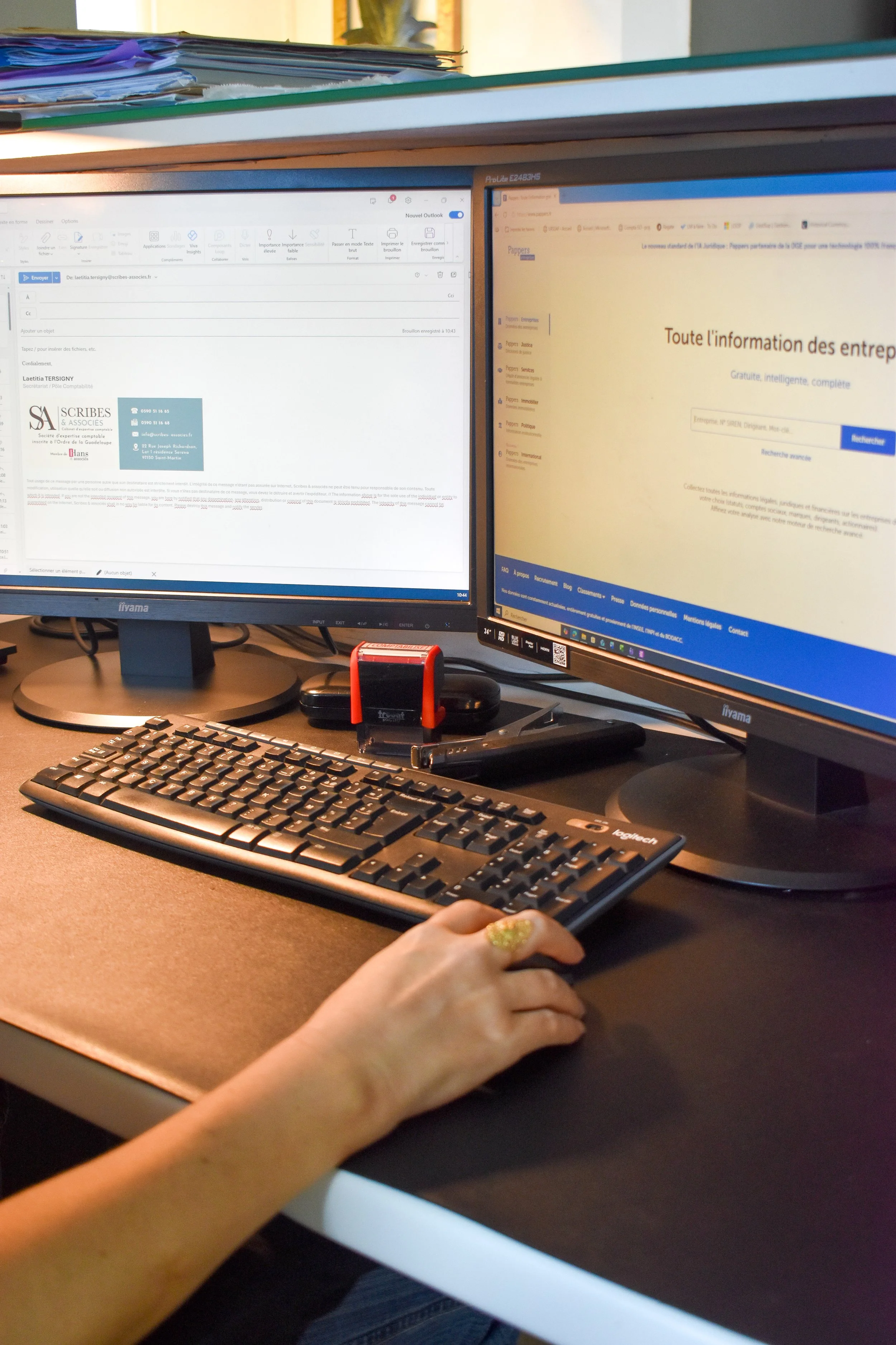 Deux écrans d'ordinateur sur un bureau avec une personne utilisant un clavier et une souris, documents, gomme, et bandeau lumineux.