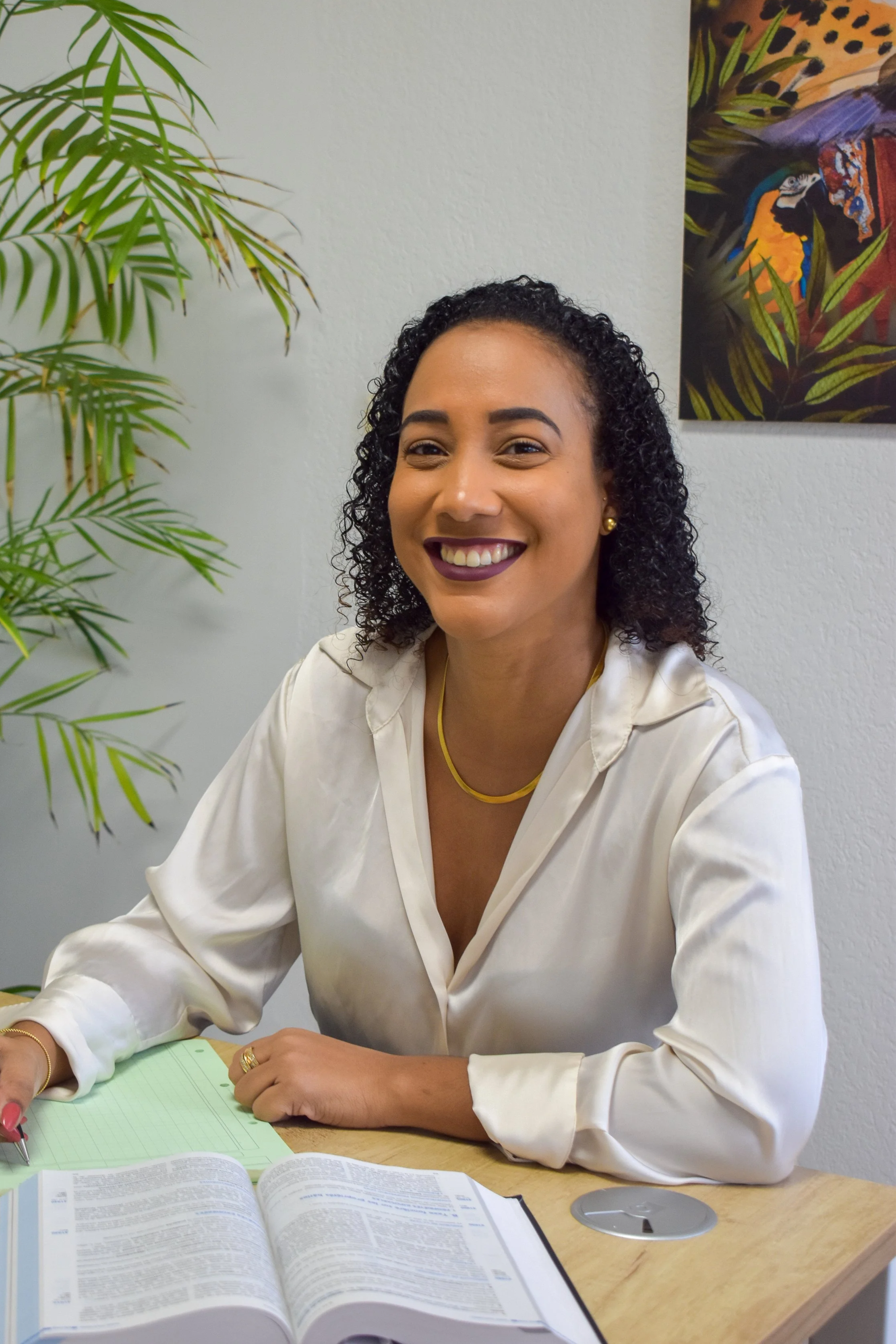 Une femme souriante avec des cheveux bouclés, portant une chemise blanche et un collier jaune, assise à un bureau avec un livre ouvert et un cahier vert, dans un bureau avec une plante verte et une peinture colorée en arrière-plan.