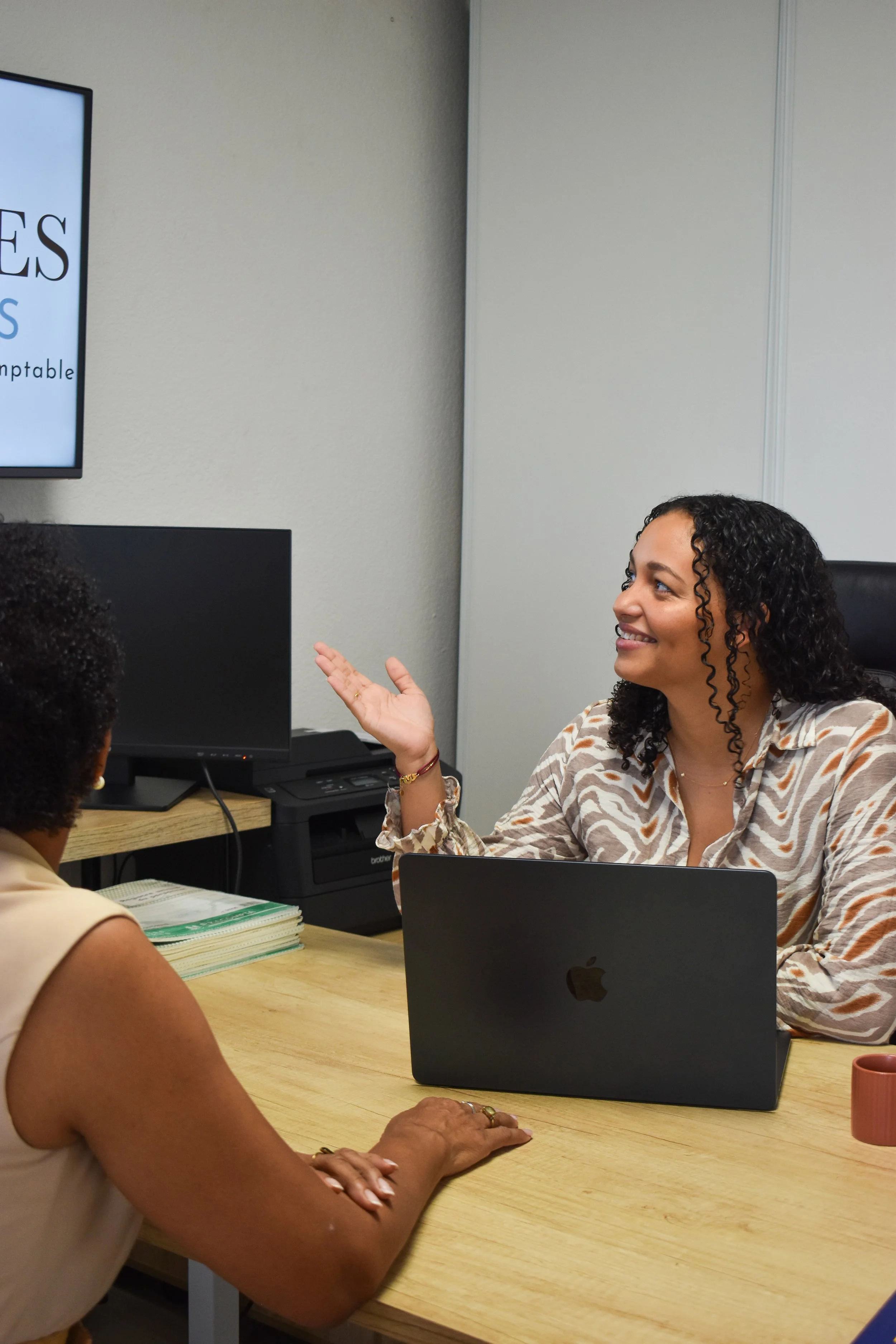 Deux femmes discutent dans un bureau, assises autour d'une table en bois avec un ordinateur portable Apple. La femme à droite, souriante, porte une chemise à motifs et a les cheveux bouclés. La femme à gauche, dont la face n'est pas visible, porte un vêtement beige. Sur le mur, un écran affiche du texte.
