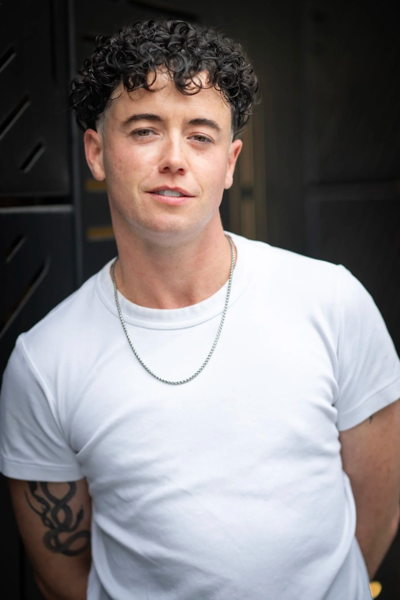A white transmasculine person with short black curly hair wears a white t shirt and chain with visible tattoos on their arms. They stare into the camera with a sly smirk.