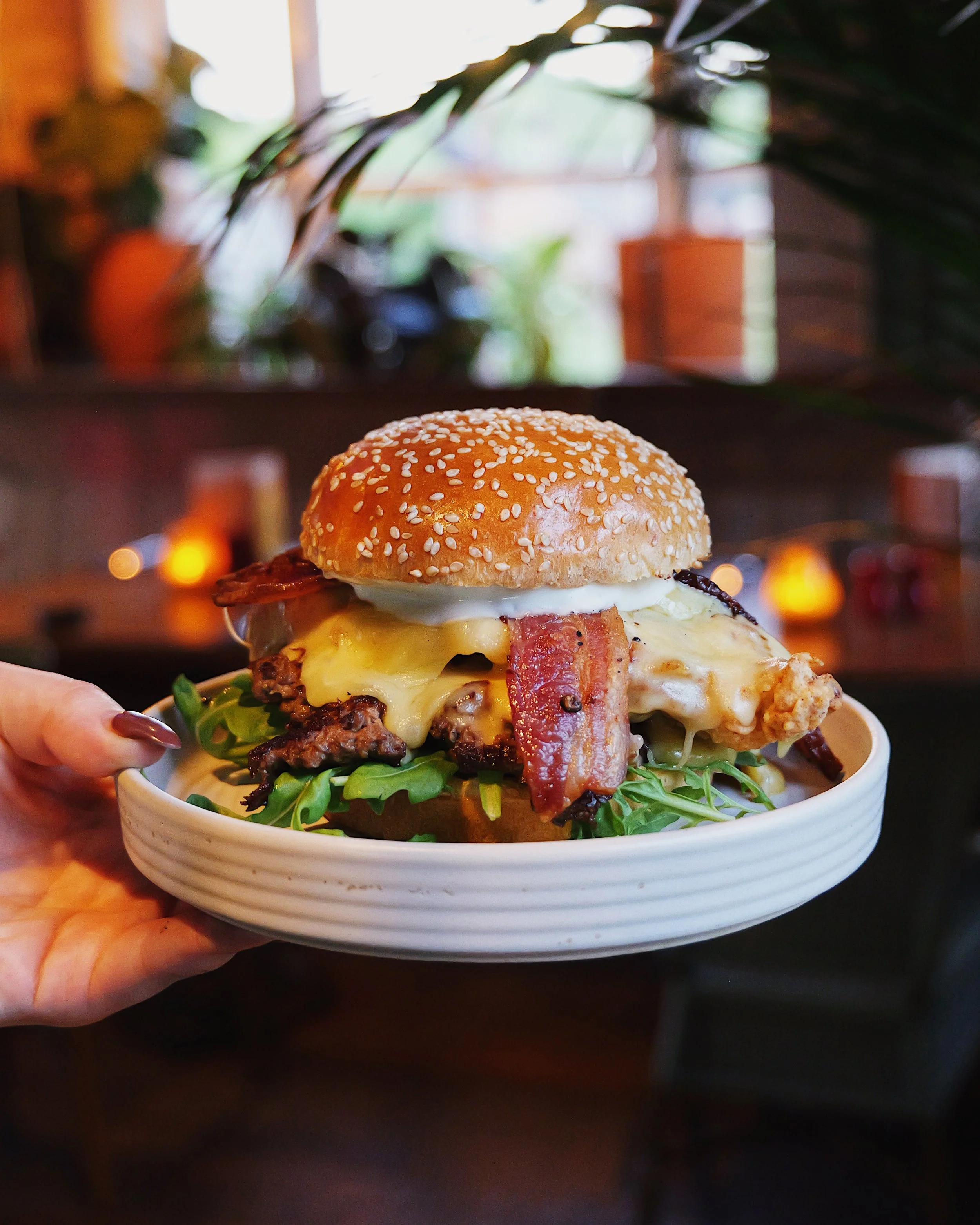 A burger topped with cheese, bacon, an egg, and sesame seed bun, served on a white plate with greens underneath, in a cozy restaurant setting.