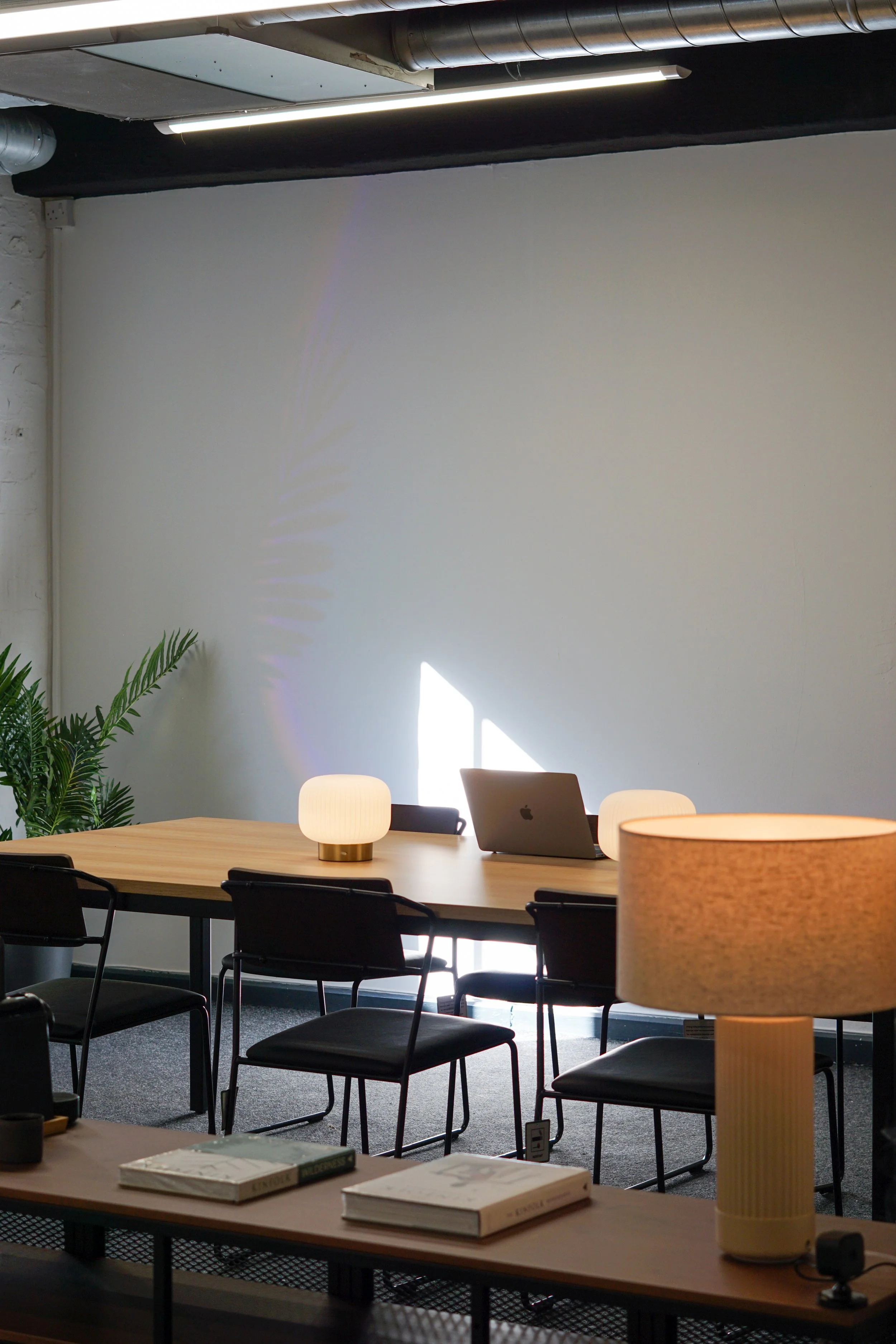 Interior of a modern conference room or office space with a wooden table, black chairs, a laptop, two table lamps, a large floor lamp, a plant, and some books, with a white wall and natural light coming in.