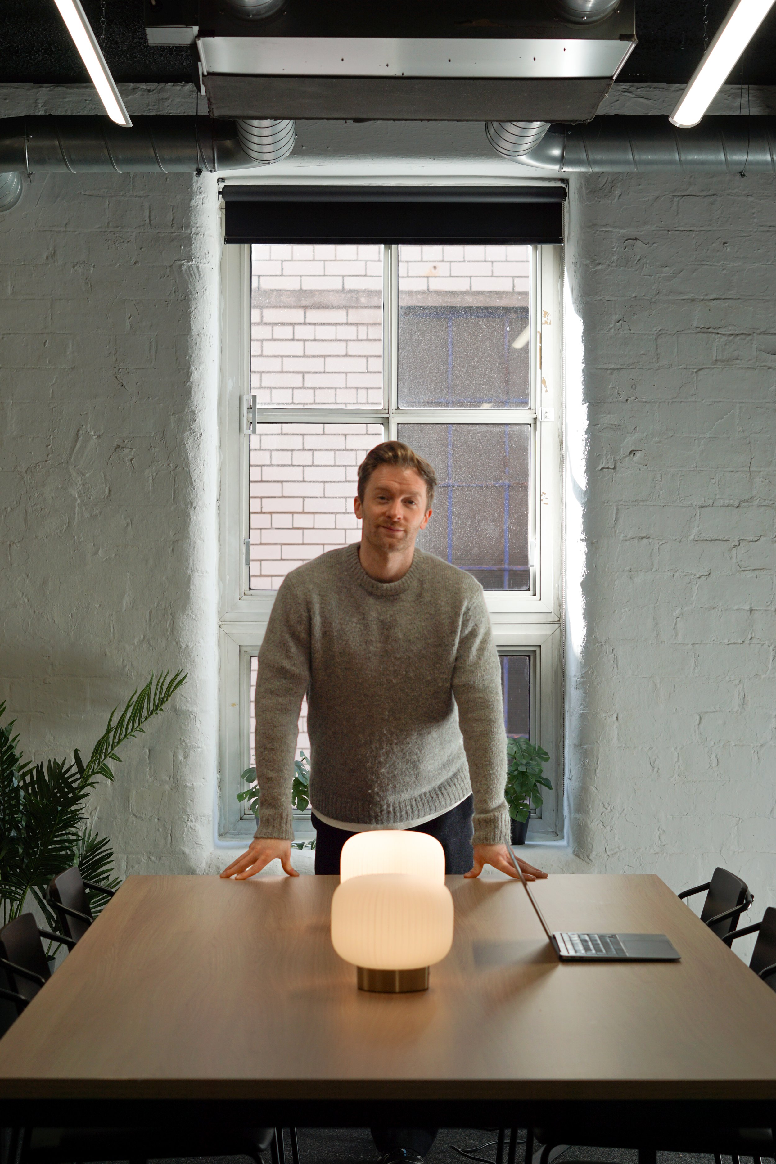 A man stands behind a conference table with a glowing lamp, in a modern office with white brick walls, a window, and plants.