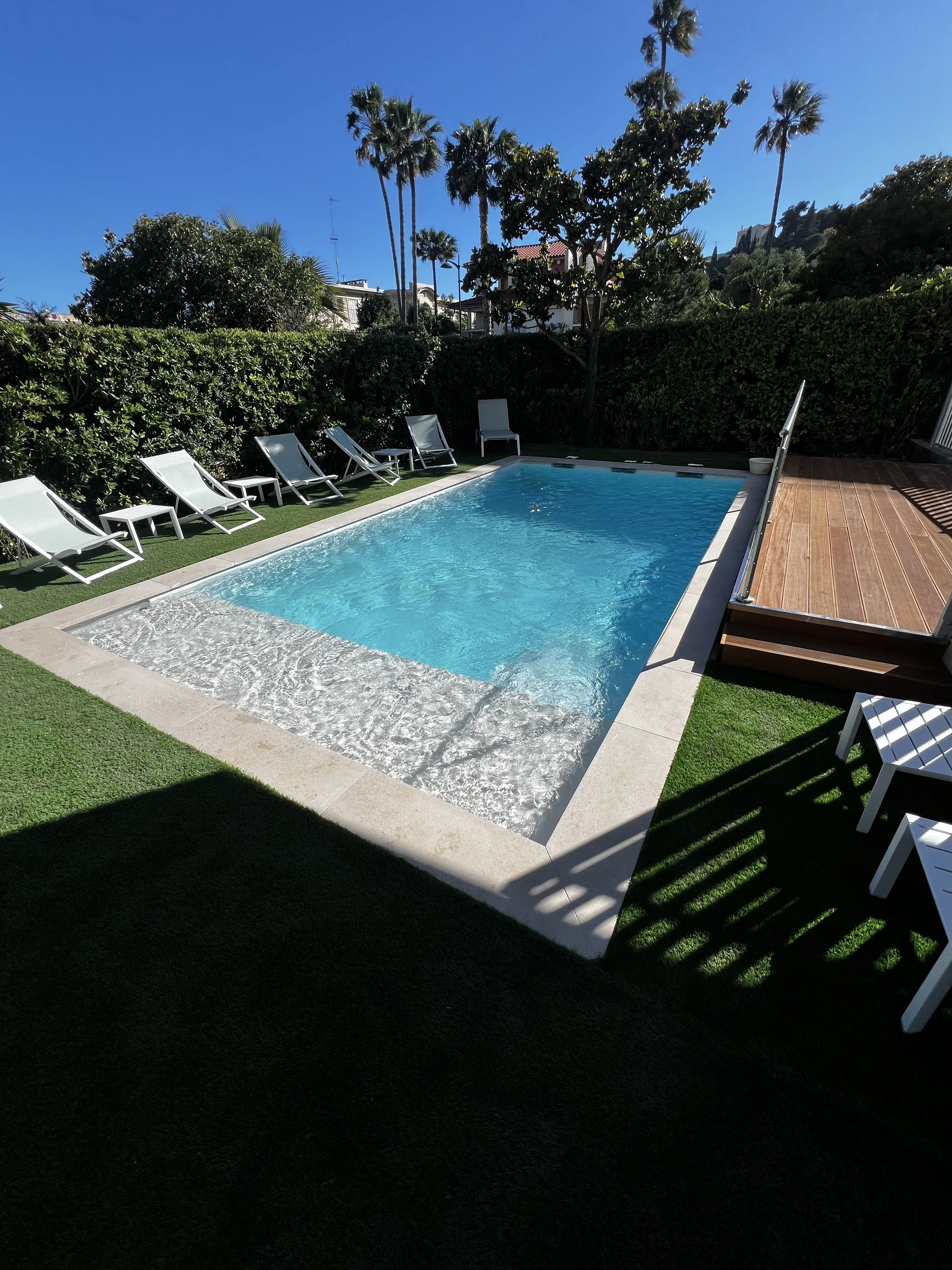 Piscine coque avec escalier d'angle, plage en dalles et terrasse bois, entourée de gazon