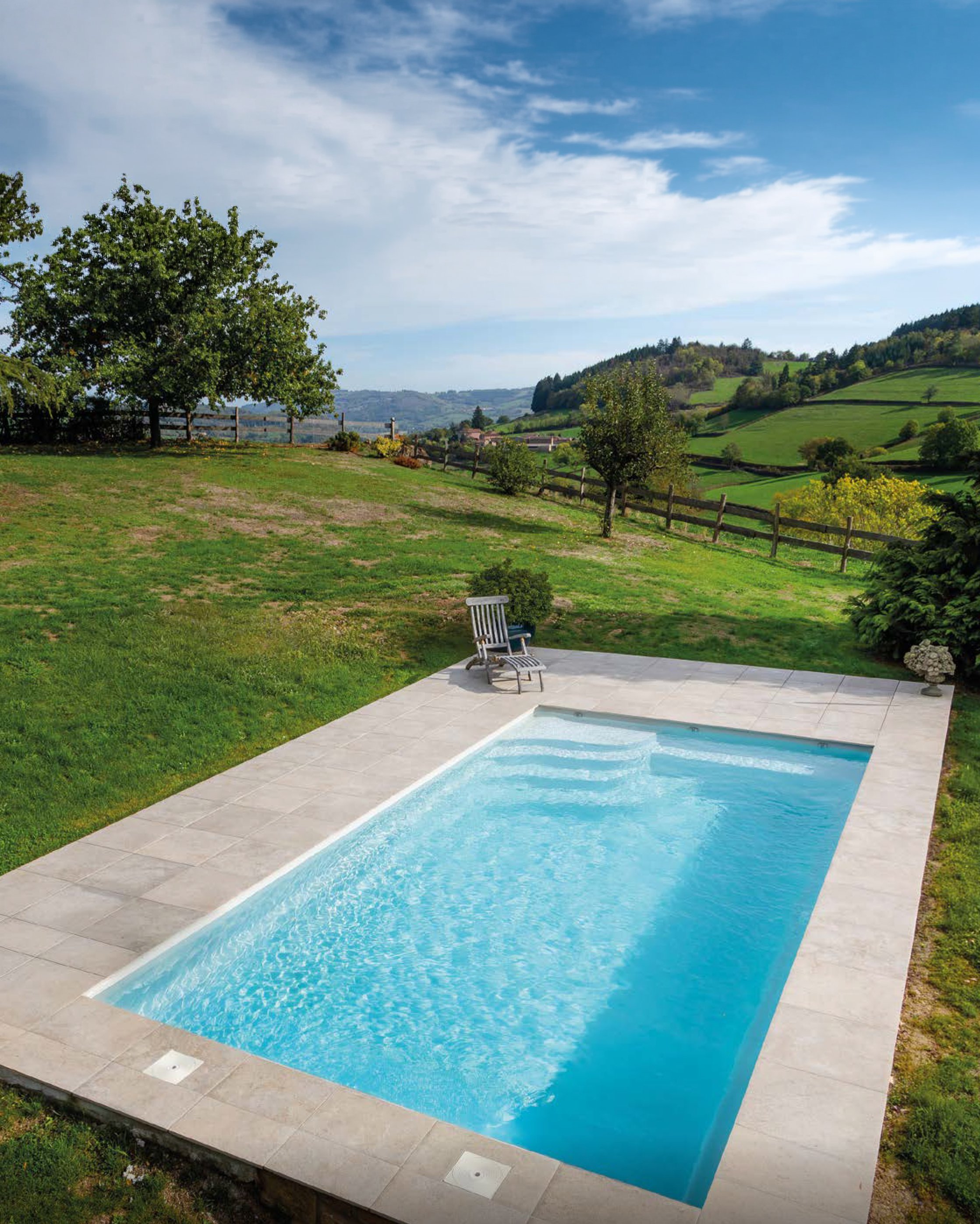 Une piscine extérieure en béton entourée d'une terrasse en carreaux, avec des chaises longues, et un paysage vallonné avec des arbres et des maisons au loin sous un ciel bleu avec quelques nuages.