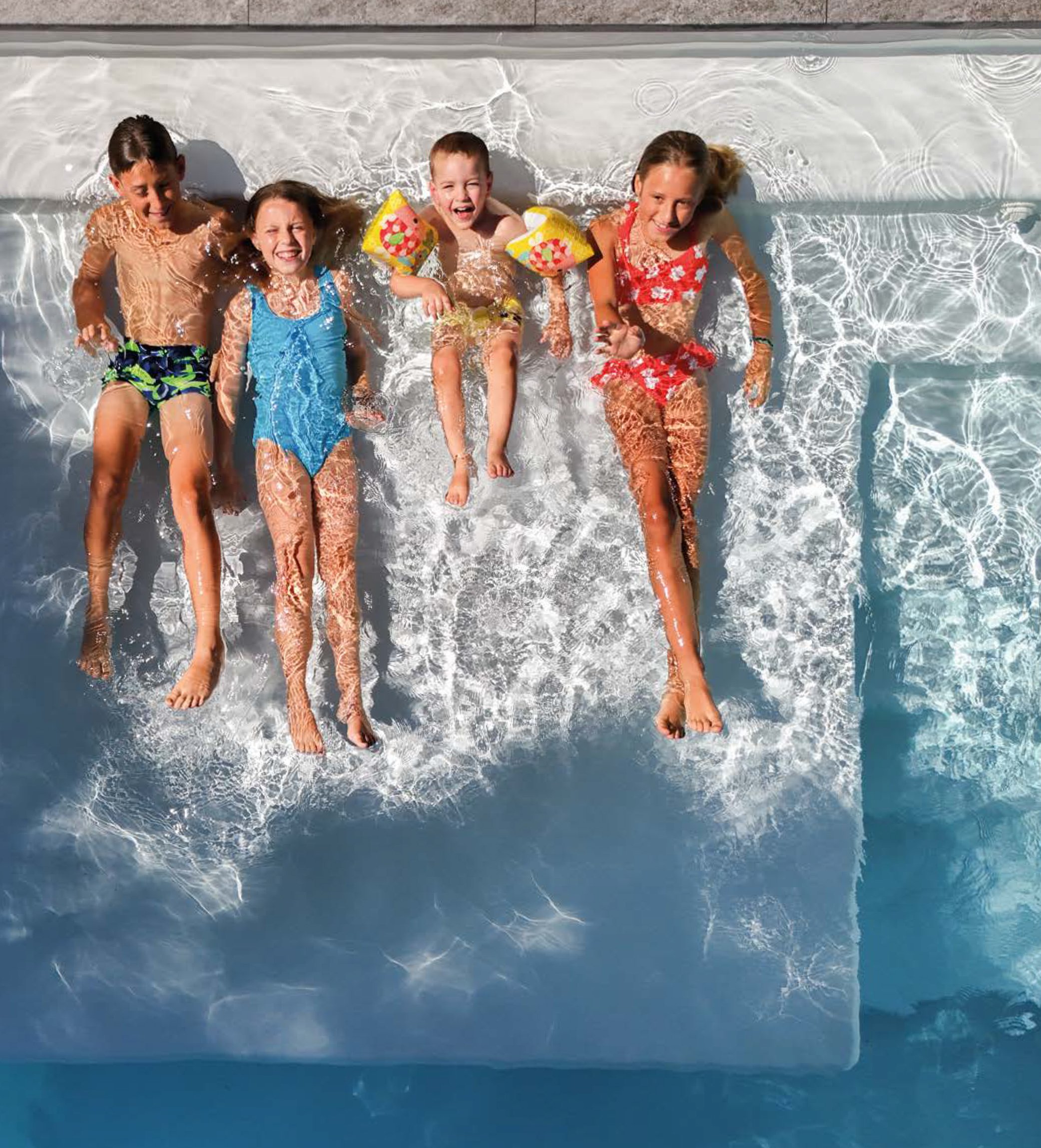 Cinq enfants souriants en maillot de bain allongés dans une piscine et jouant avec des brassards gonflables jaunes, vue du dessus.