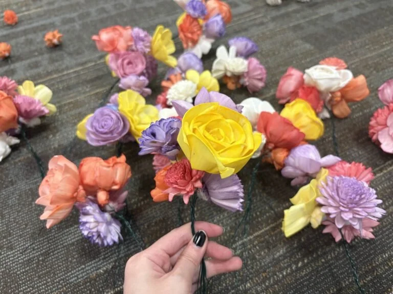 Photo of a Remnants team member creating and assembling the decorative flowers for the Remnants trellis. The flowers are being held by the team member and the flowers are purple, red, orange, yellow, and white. Photo: Remnants Team.