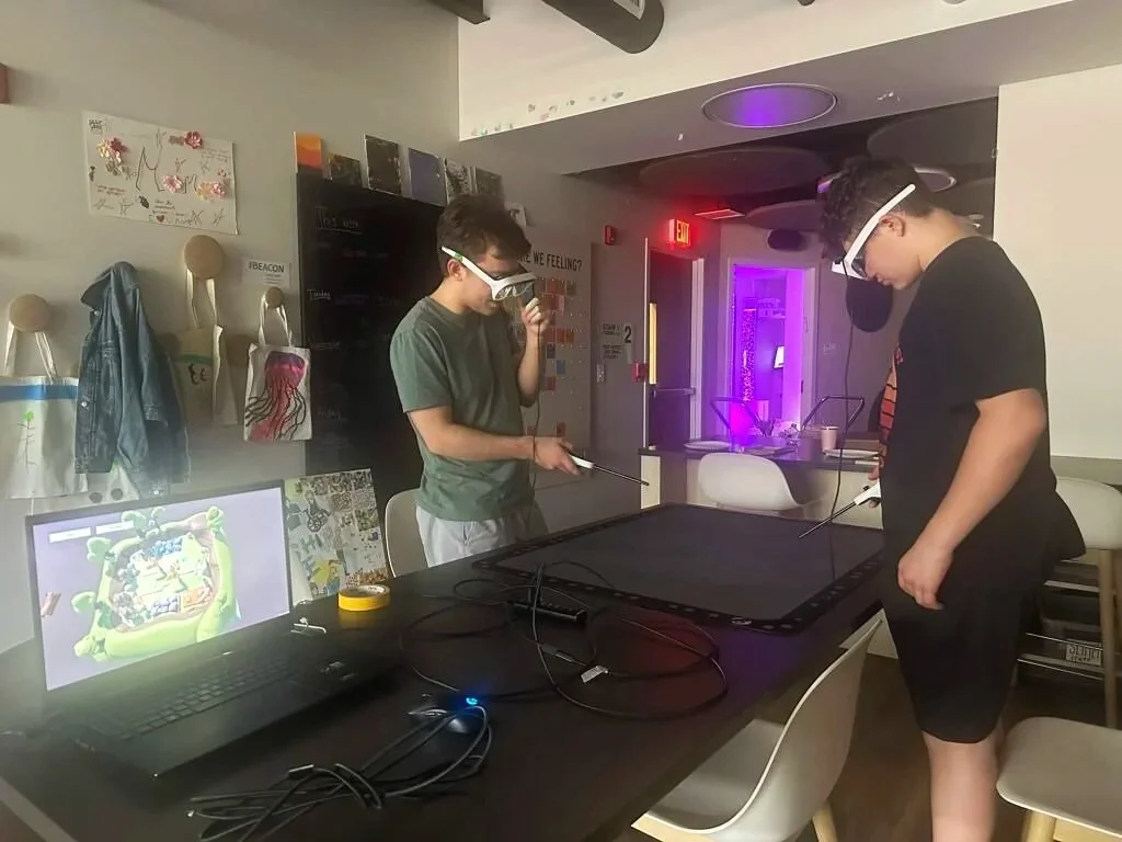 Photo of two students wearing augmented reality glasses and using "magic wands" around the Tilt Five playmat, an augmented reality collaborative game space. Source: It Takes 2 Alices.