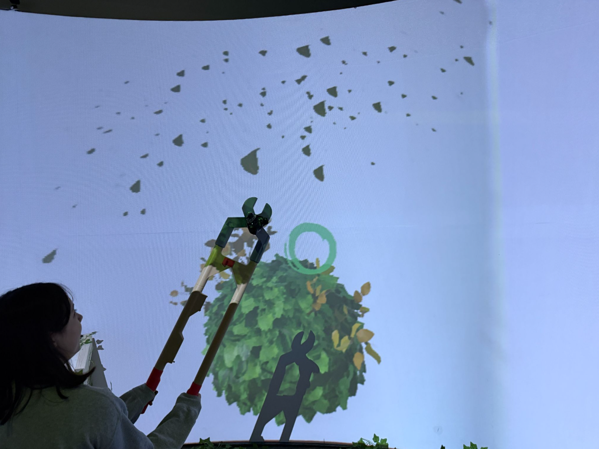 Photo of a guest interacting with Remnants using a physical prop, a pruning shear, and watching the resulting action on the curved CAVERN screen. The screen shows a tree with leaves being cut. Photo: Remnants Team.