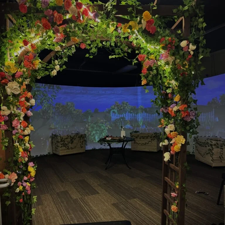Photo showing the entryway to Remnants in the Carnegie Mellon University Entertainment Technology Center CAVERN space. The guest walks under a garden trellis decorated with flowers painted red, pink, yellow, white, and purple. Photo: Remnants Team.