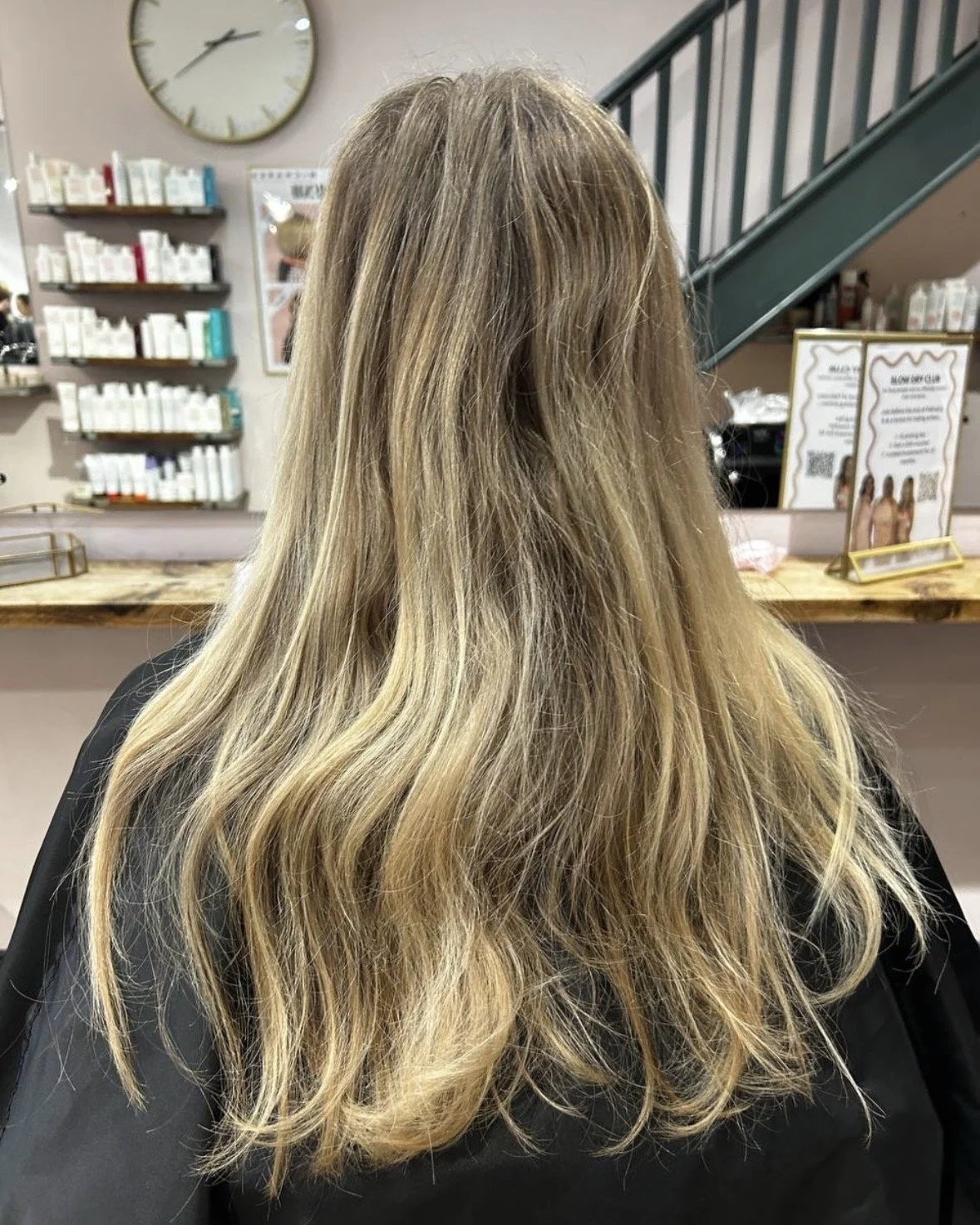 Back of a person's head with long, wavy blonde hair in a salon, with shelves of hair products and a large wall clock in the background.