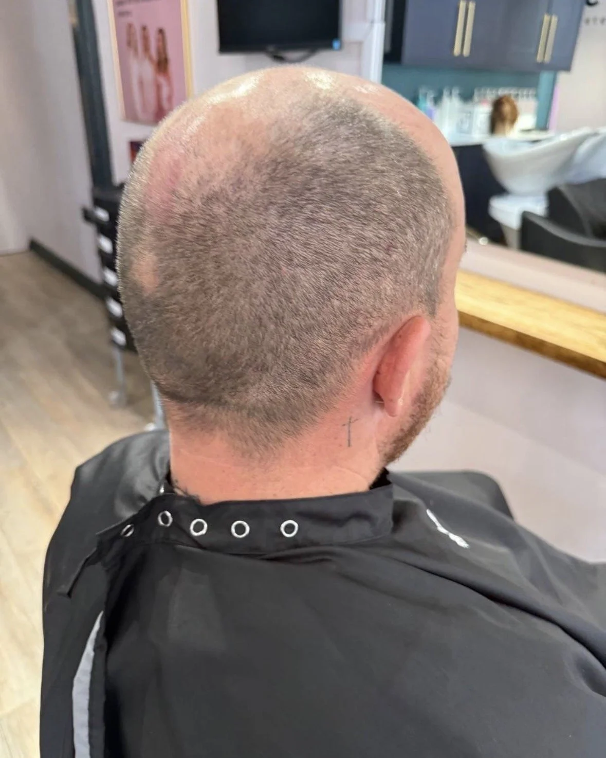 Back of a person's head with a freshly shaved hairstyle in a salon, with a small cross tattoo visible on the neck.