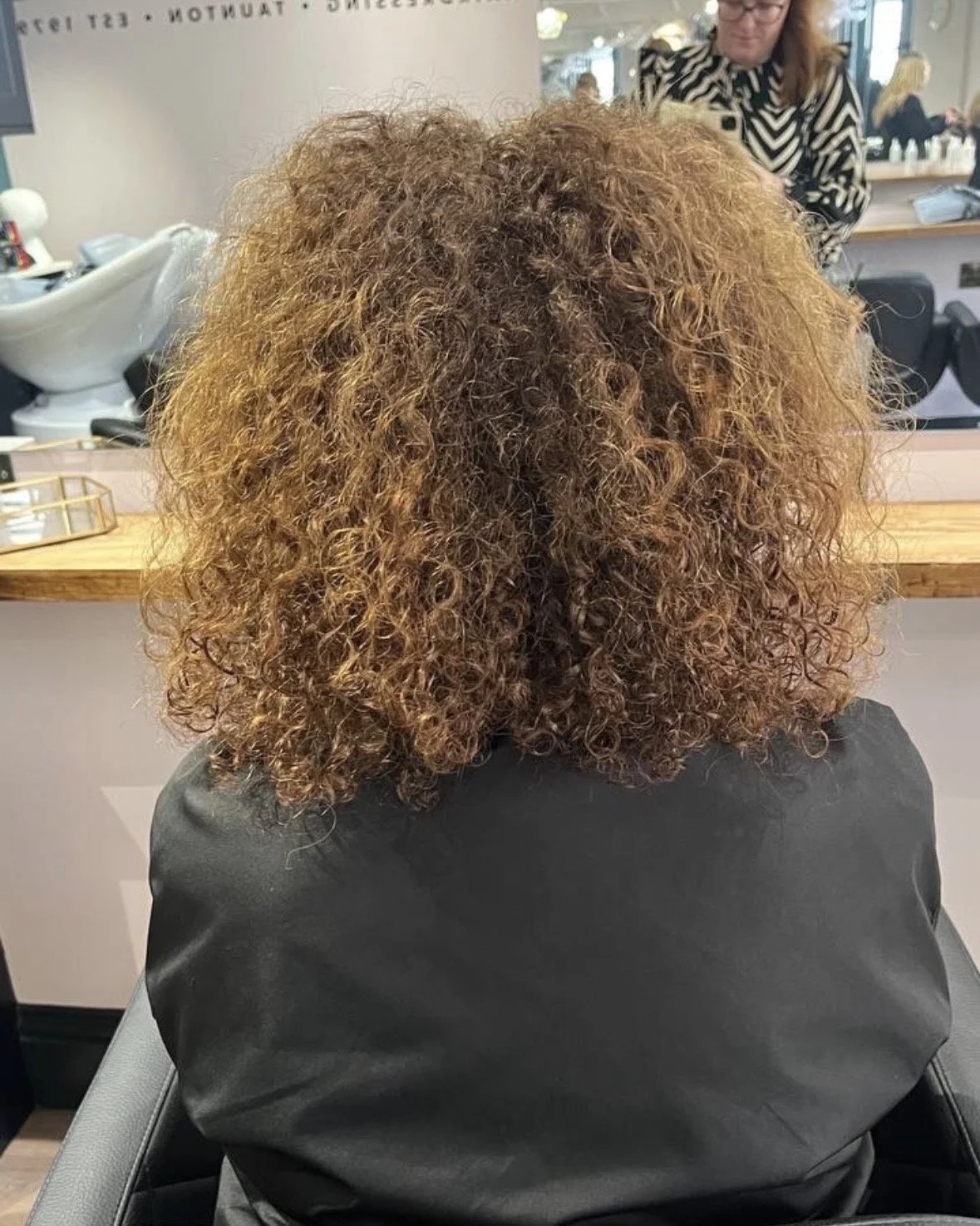 Back view of a woman with shoulder-length, curly, brown hair sitting in a salon chair at a hair salon.