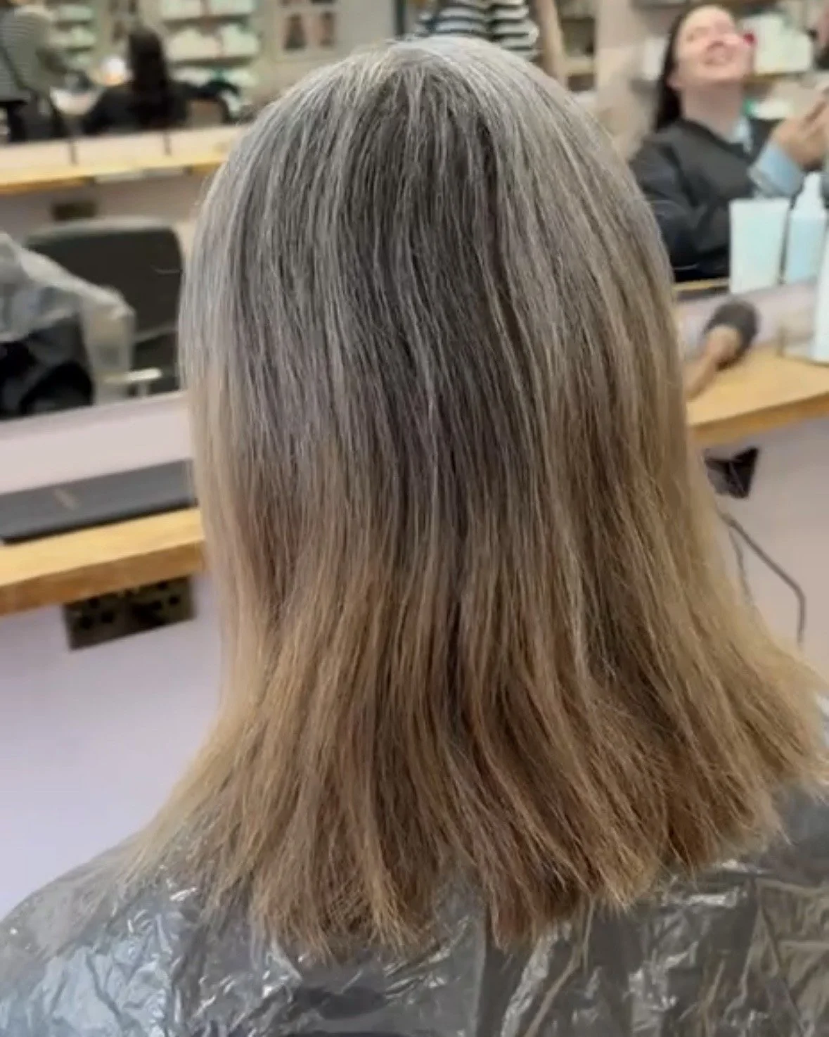 Back view of a woman with shoulder-length, blonde hair in a hair salon, sitting in a chair with a protective cape around her shoulders. The background shows other customers and salon equipment.