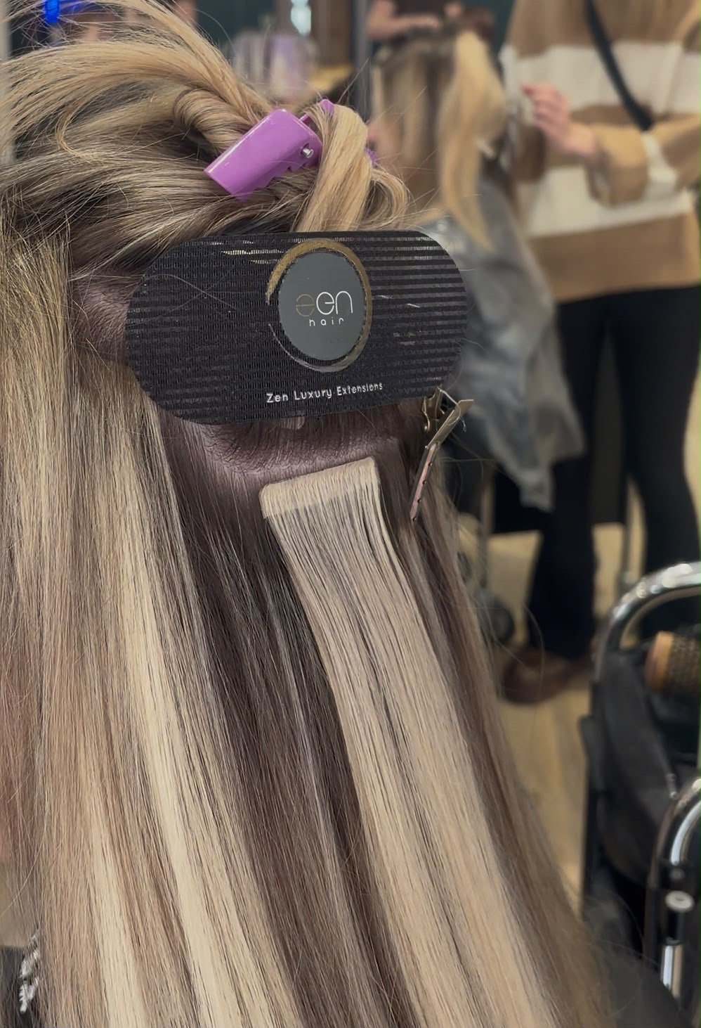 A person getting hair extensions in a salon, with blonde hair, a purple hair clip, and a black label indicating "Zen Luxury Extensions".
