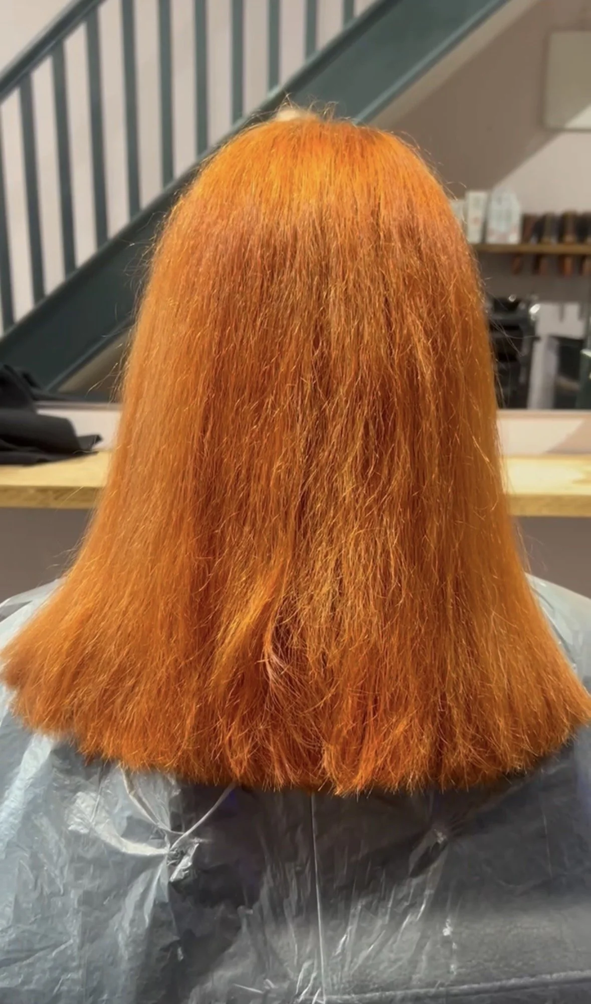 Back view of a person with long, straight, bright orange hair. The person is seated on a black salon chair covered with a plastic cape, in a salon setting with a staircase and shelves in the background.