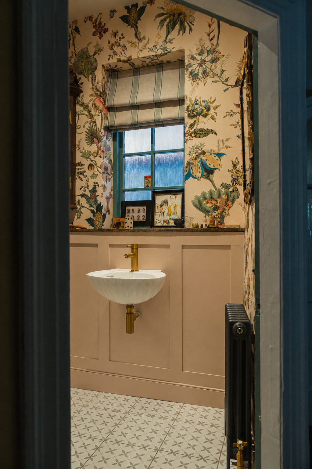 View into a richly layered downstairs powder room featuring botanical wallpaper, brass fittings, bespoke panelling, and a sculptural sink design.