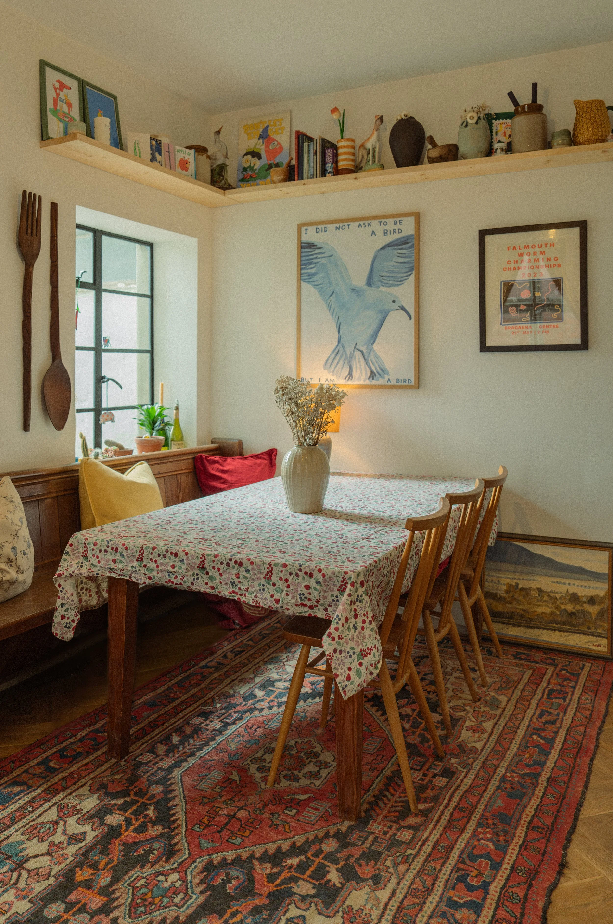 A dining room with a wooden table covered in a floral tablecloth, surrounded by wooden chairs. A large Persian rug covers the floor. The wall displays framed artwork, including a painting of a bird and a framed poster for a worm charming championship. Decorative shelves hold books and pottery, and a window with small plant decorations is visible. A vase with dried flowers is on the table.