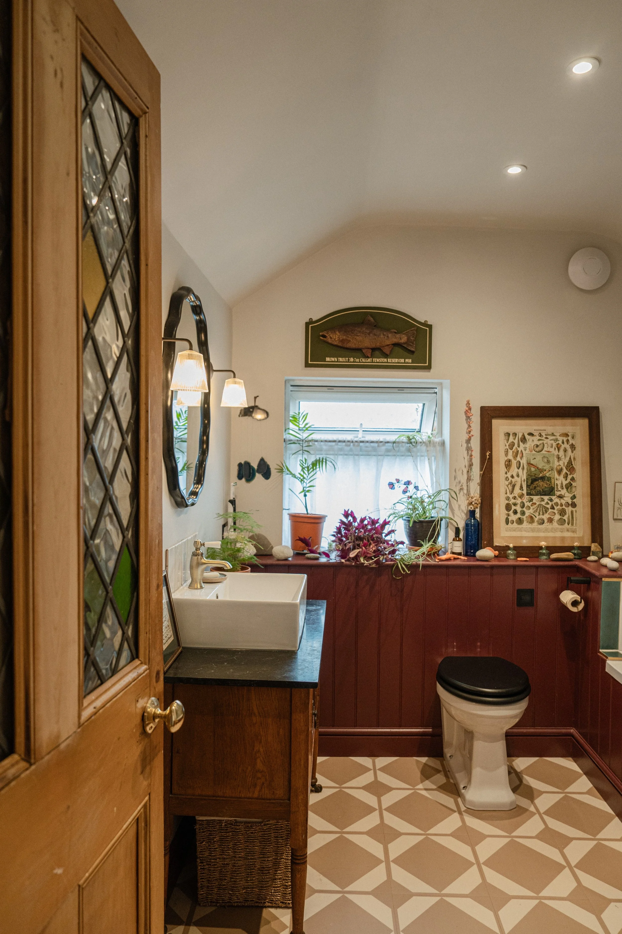 View of a bathroom seen through an open wooden door with a diamond pattern glass panel. The bathroom has a white toilet with a black seat, a black countertop with a white square sink, and wall-mounted lights above. A window above the sink lets in natural light, revealing potted plants and decorative items on the counter. There are framed artworks and a mounted fish plaque on the walls, with a patterned tile floor.