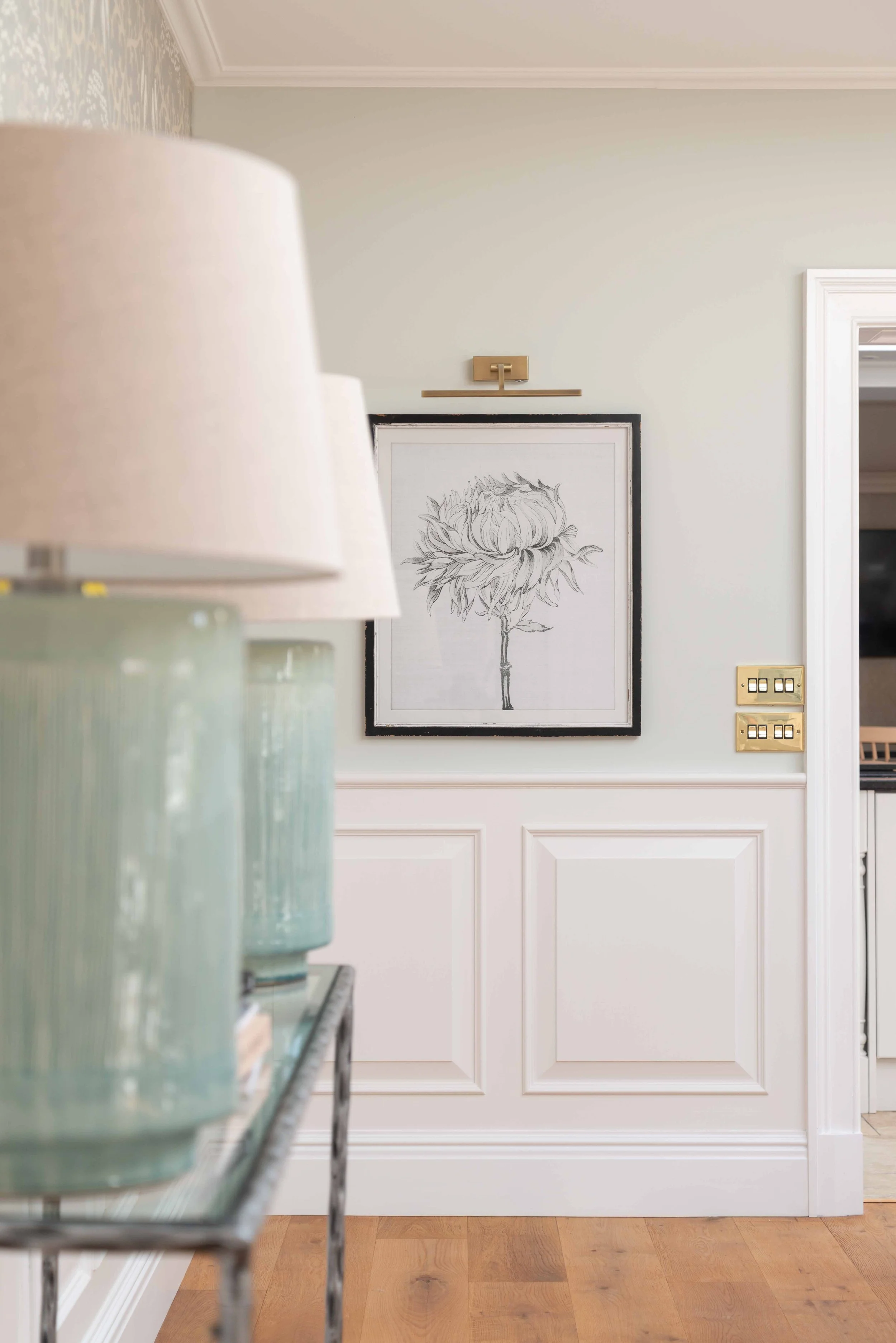 A hallway with a framed black and white floral drawing on the wall, a white door with decorative panels, and gold switch plates.