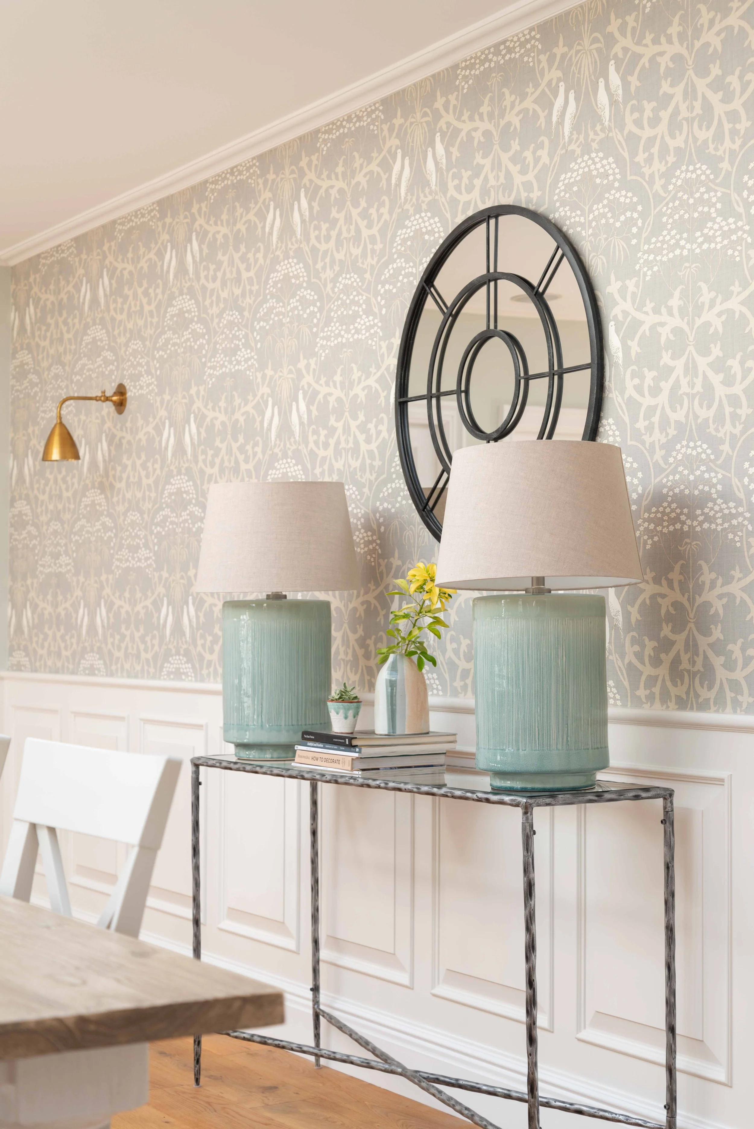 Interior room with a patterned wallpaper featuring birds and floral designs. A large round black metal wall decor hangs on the wall. Two tall light green ceramic vases with beige lampshades are placed on a metal console table. A small plant and a few books are also on the table. There is a white chair partially visible in the foreground and a wooden surface on the left side.