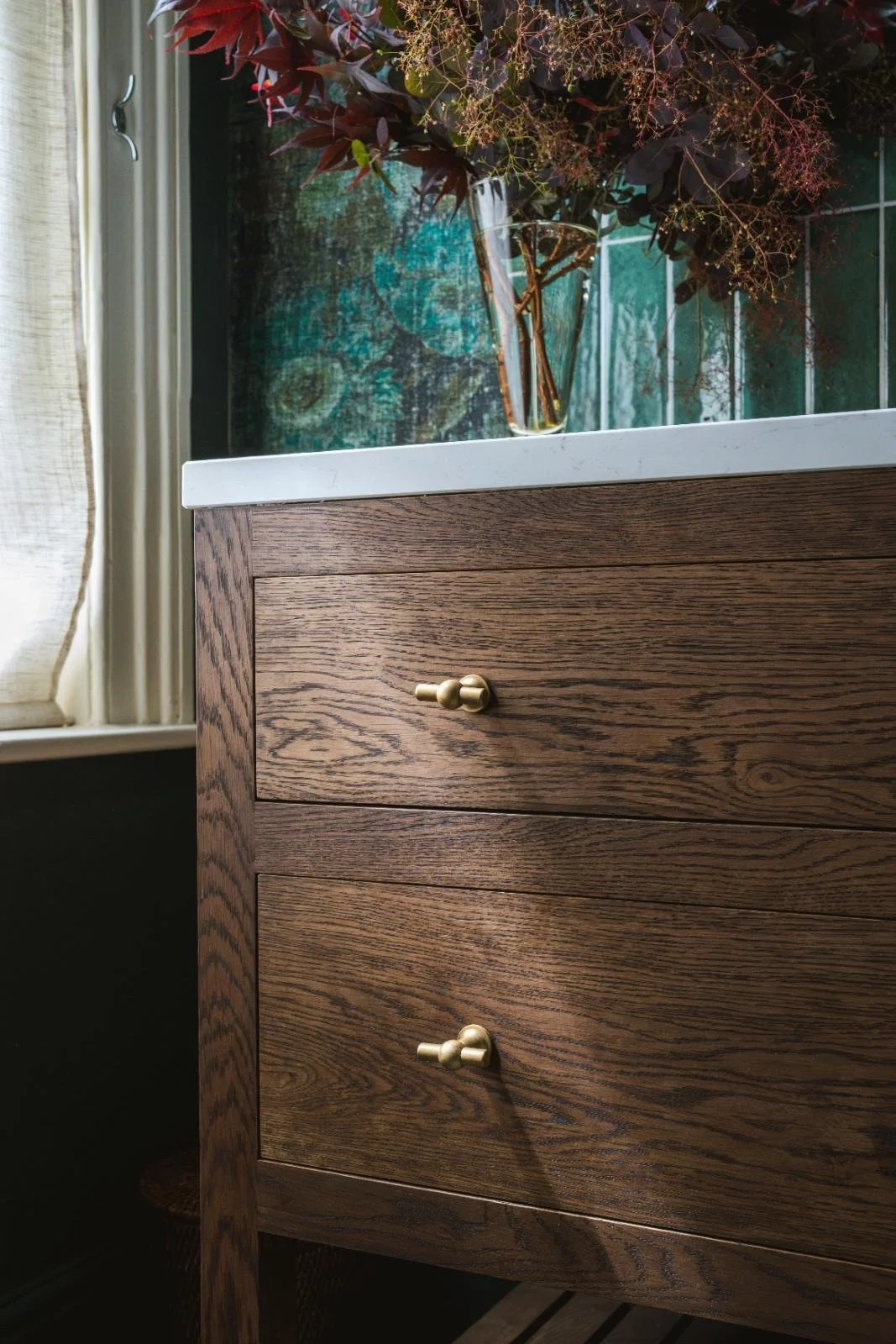 A wooden dresser with three drawers and small gold knobs, next to a corner of a window with a beige curtain. On top of the dresser, there is a glass vase with dried flowers and greenery, positioned in front of a colorful, abstract painting or tapestry.