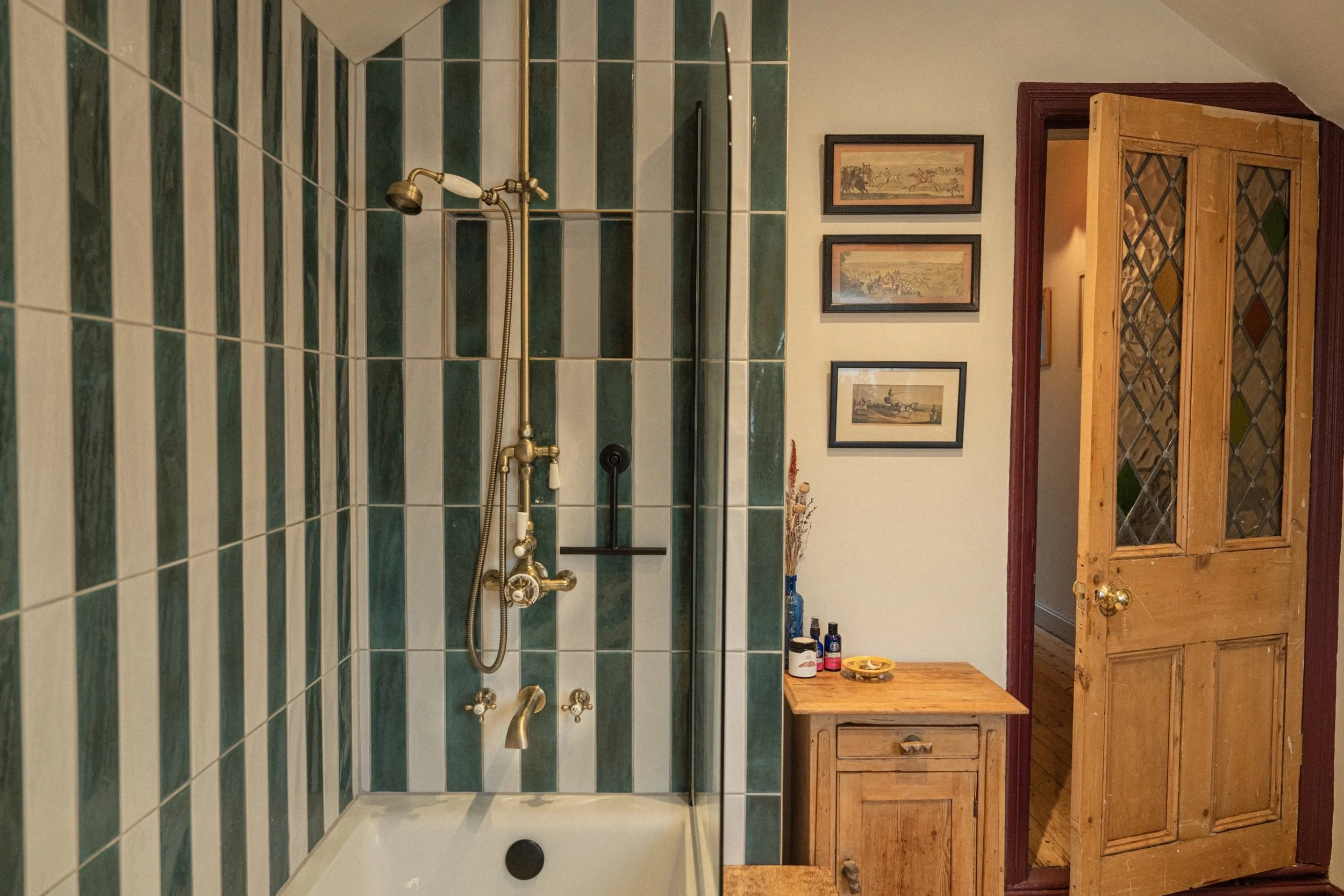 Edwardian townhouse bathroom with striped green wall tiles, brass shower fittings, original timber door, and collected vintage styling.