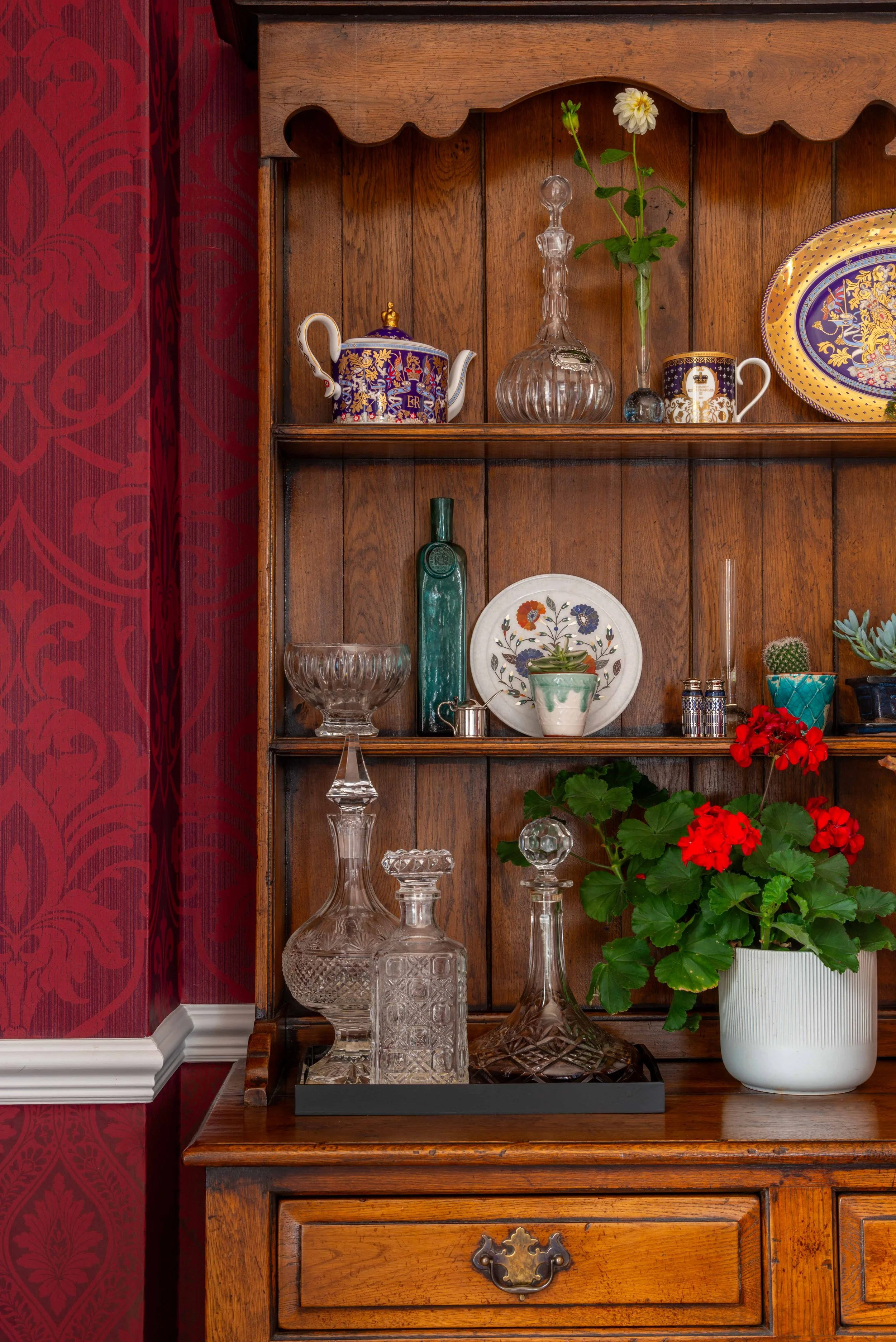 A wooden hutch shelf with decorative glassware, porcelain teapots, a floral plate, a potted plant with red flowers, and a red damask curtain to the side.