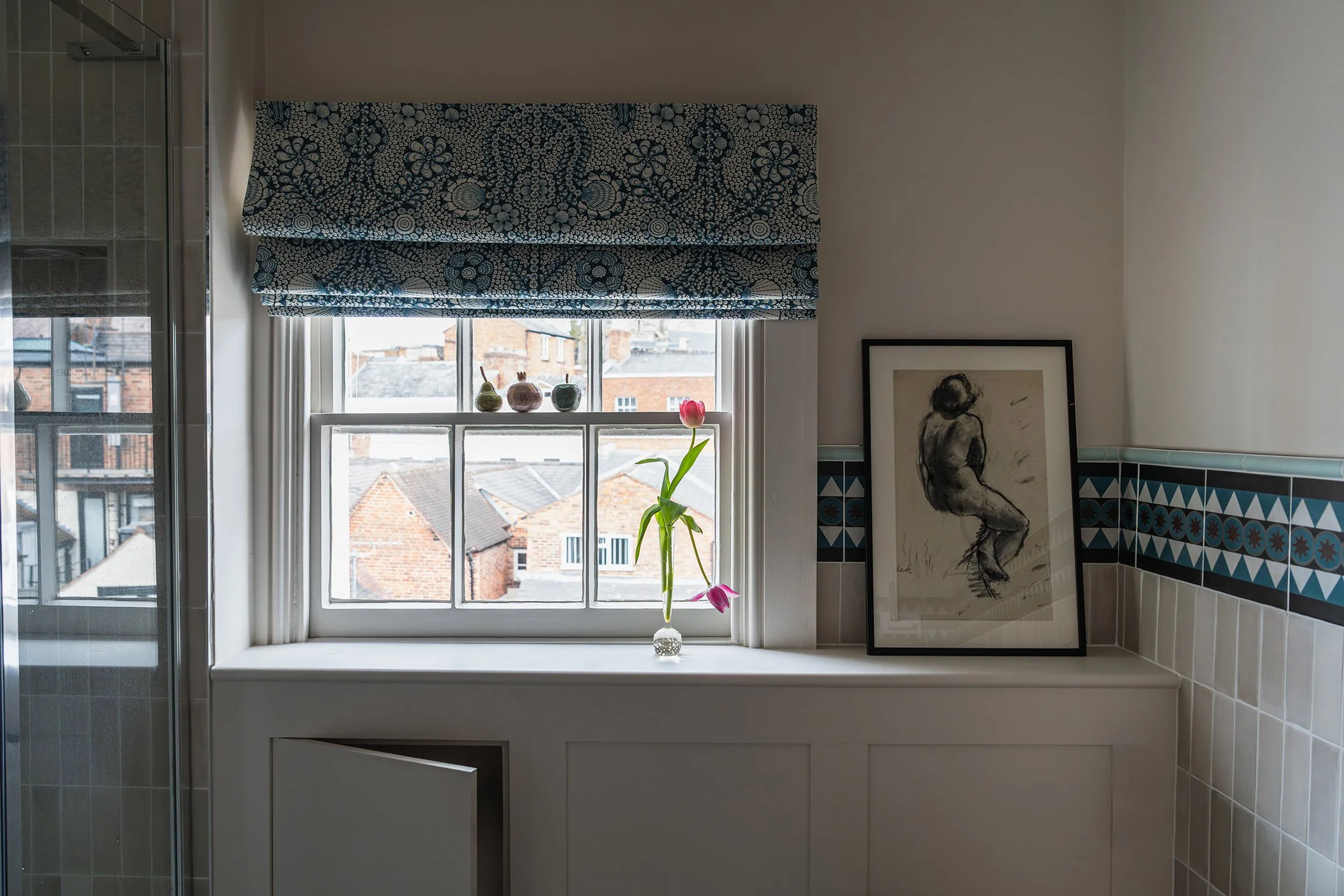 Townhouse bathroom with decorative tile border, botanical Roman blind, framed artwork, and soft natural light creating a calm retreat.