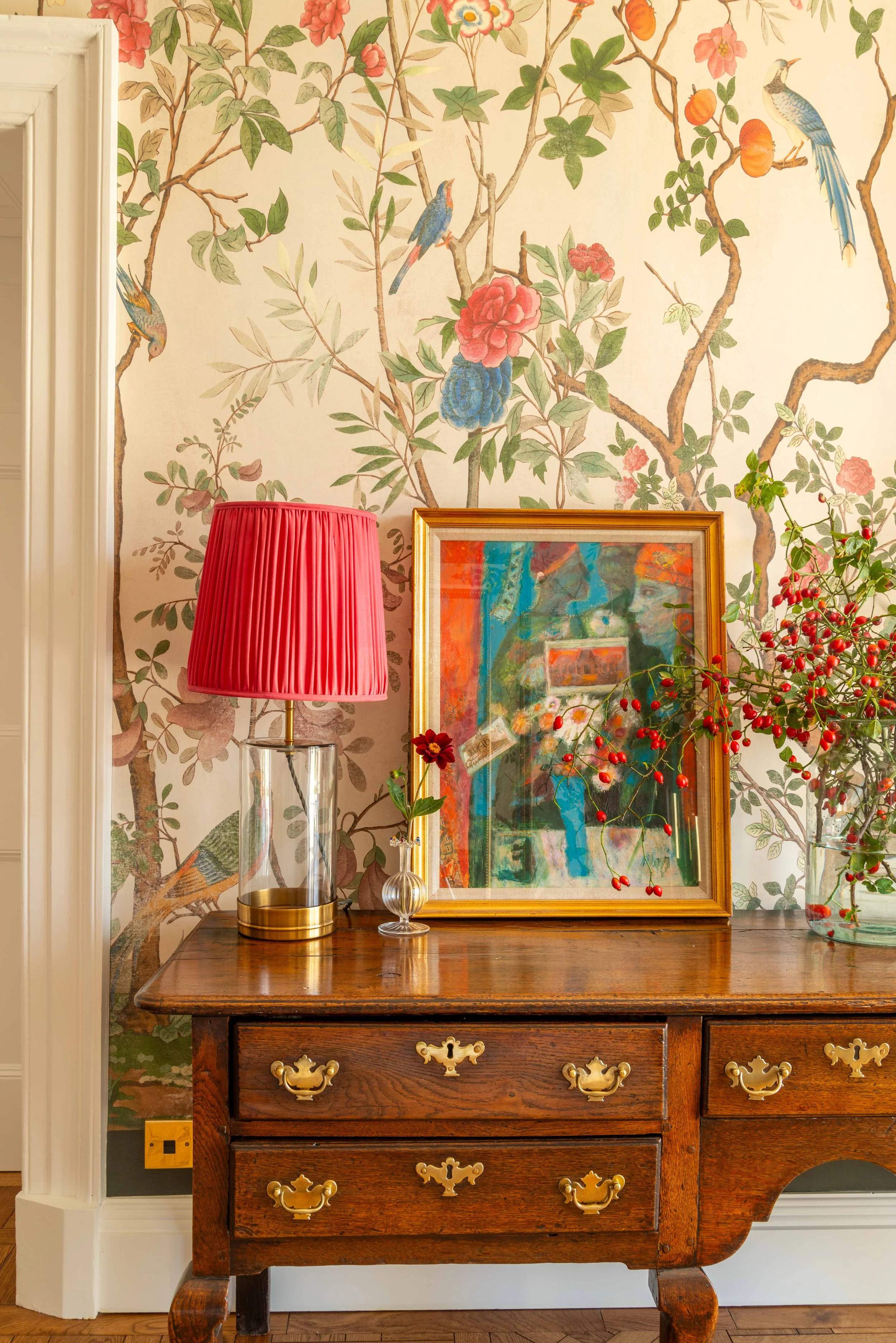 Antique chest of drawers styled with layered artwork, floral wallpaper, and rich red table lamp in an elegant Georgian hallway interior.