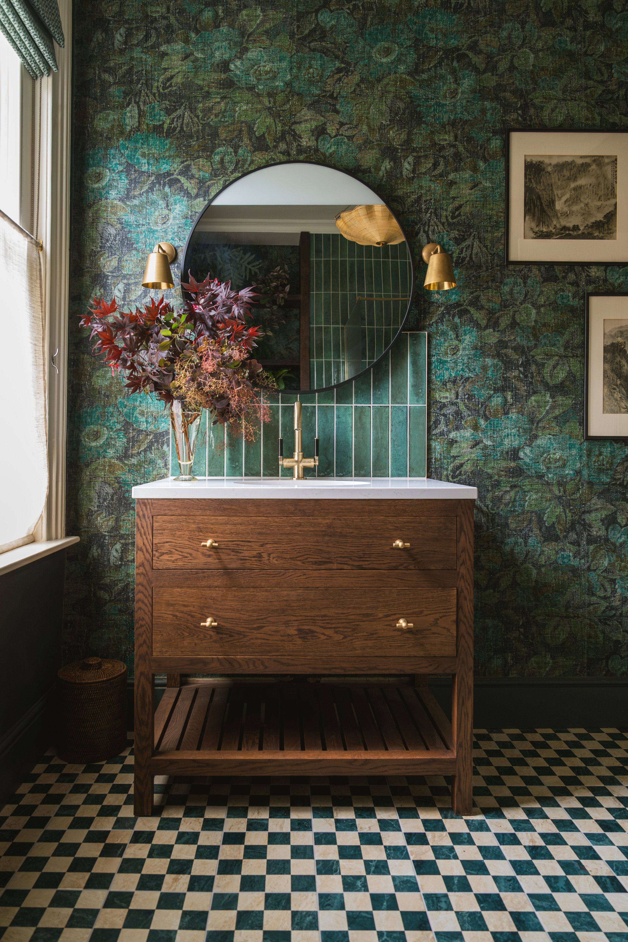 Bathroom with a wooden vanity, round mirror, green tiled backsplash, and a black and white checkered floor. There is a vase with dark red and purple leaves and a framed artwork on the wall.